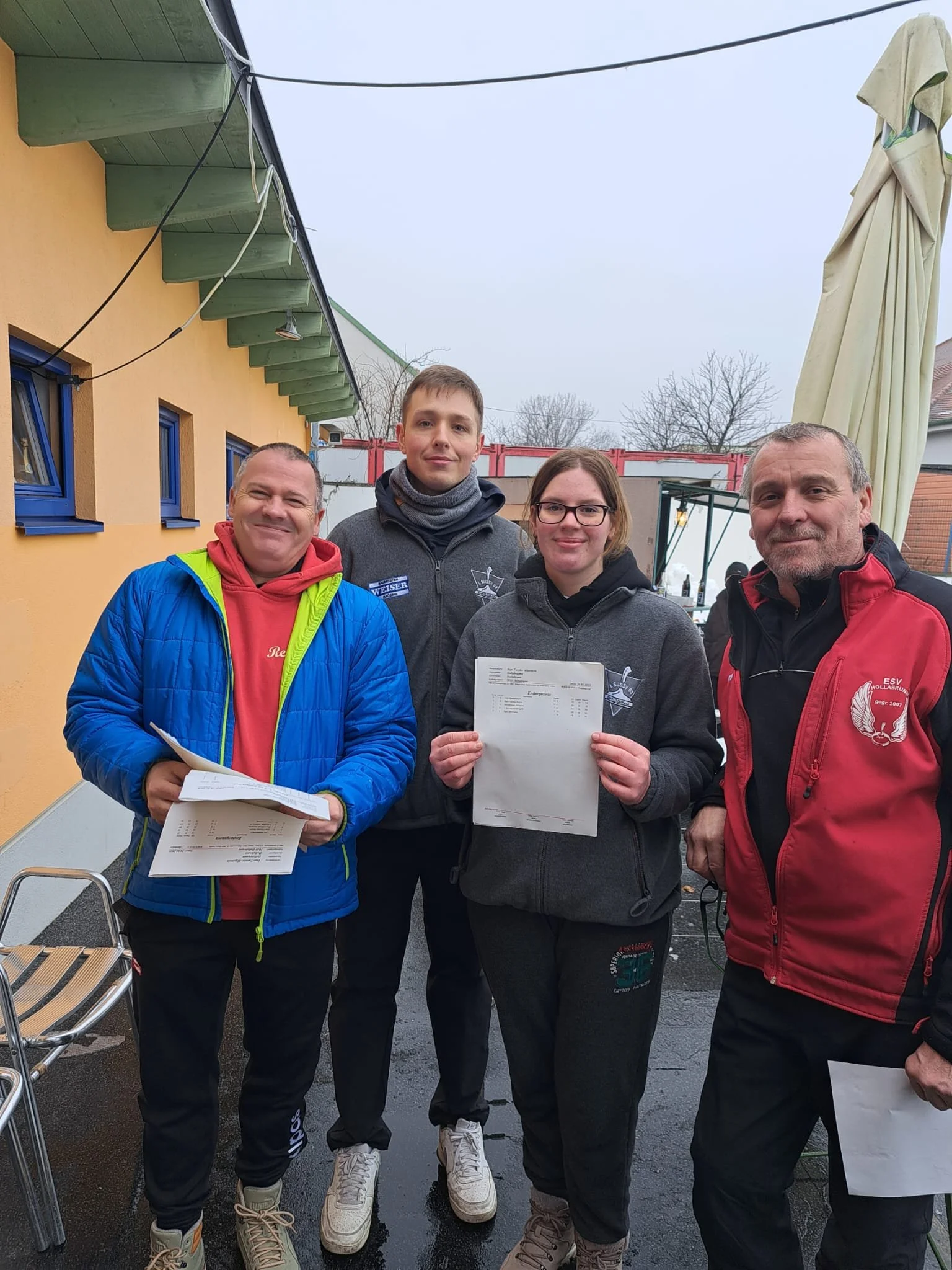 Vier Menschen, zwei Männer und zwei Frauen, stehen zusammen vor einem gelben Gebäude mit blauen Fenstern. Sie halten Papiere in den Händen und posieren für ein Foto. Es ist draußen, es sieht bewölkt aus, und im Hintergrund sind Bäume und eine geschlossene große Sonnenschirm zu sehen.