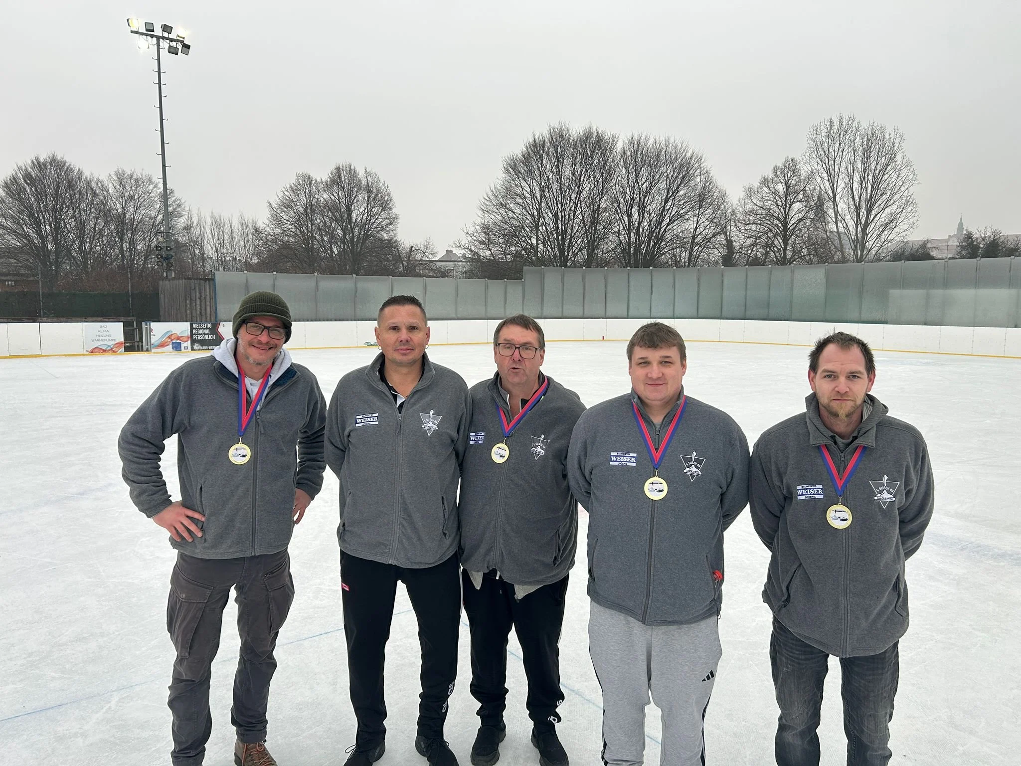 Fünf Männer auf einer Eisbahn, jede mit einer Goldmedaille um den Hals, tragen graue Jacken mit Logos, im Hintergrund Bäume und eine umzäunte Eisfläche bei bewölktem Himmel.
