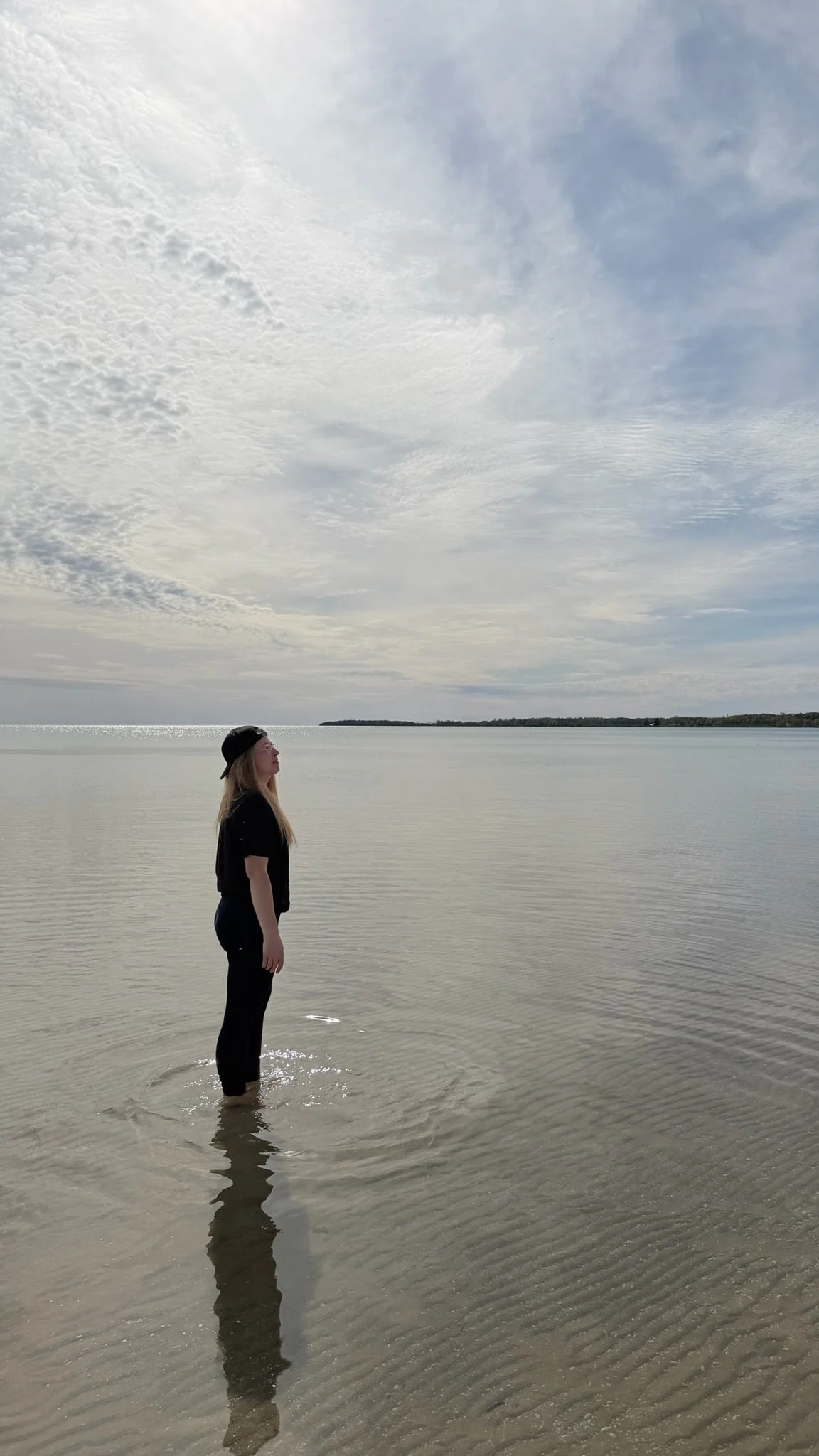 A woman standing in calm water at the edge of a lake or sea, wearing black clothing and a black cap, looking to the left. The water reflects her silhouette and the sky is cloudy with patches of blue.