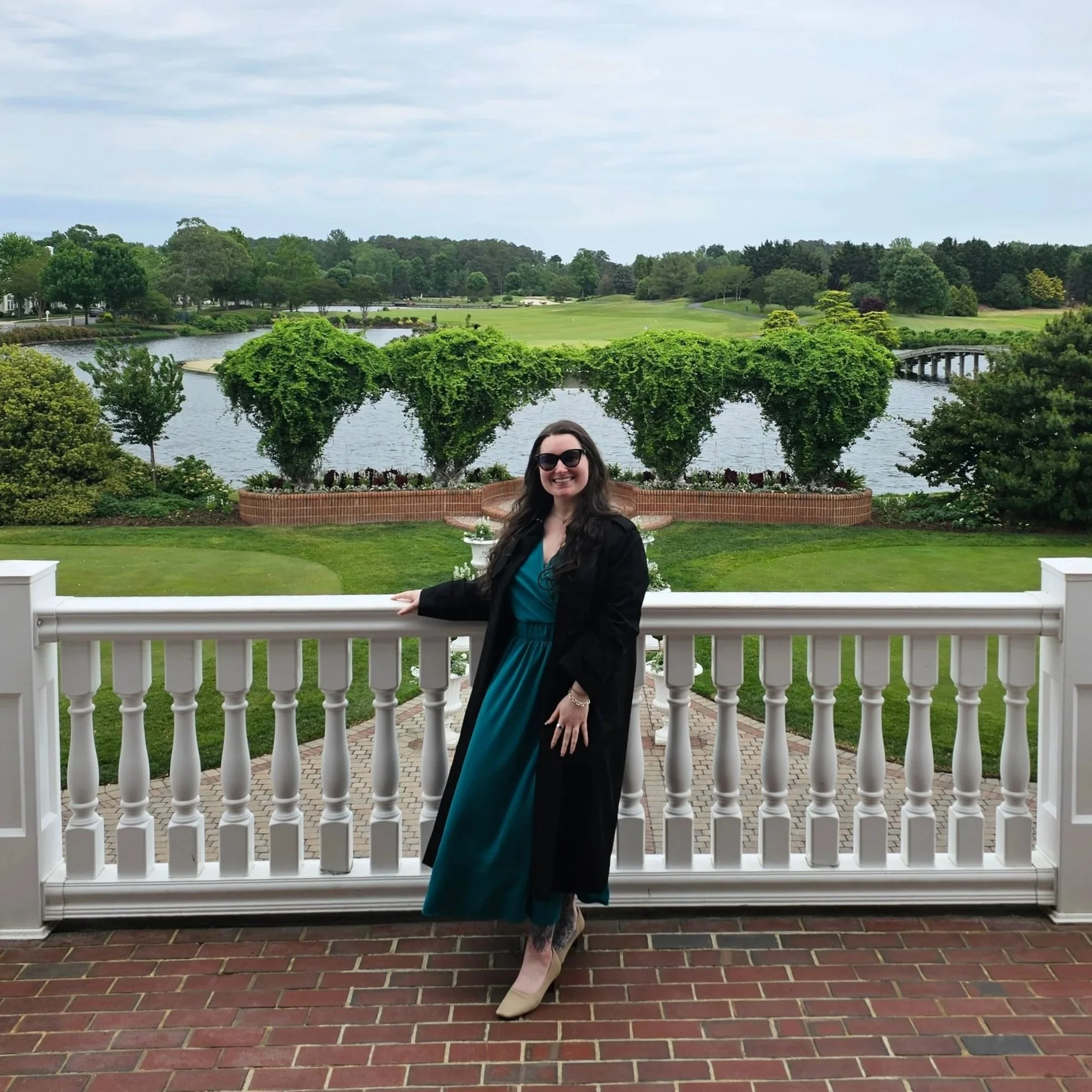 A woman wearing sunglasses, a teal dress, and a black jacket standing on a brick patio next to a white railing, smiling for the camera. Behind her, there are landscaped gardens, a pond, a bridge, and a lush green golf course with trees and hills under an overcast sky.