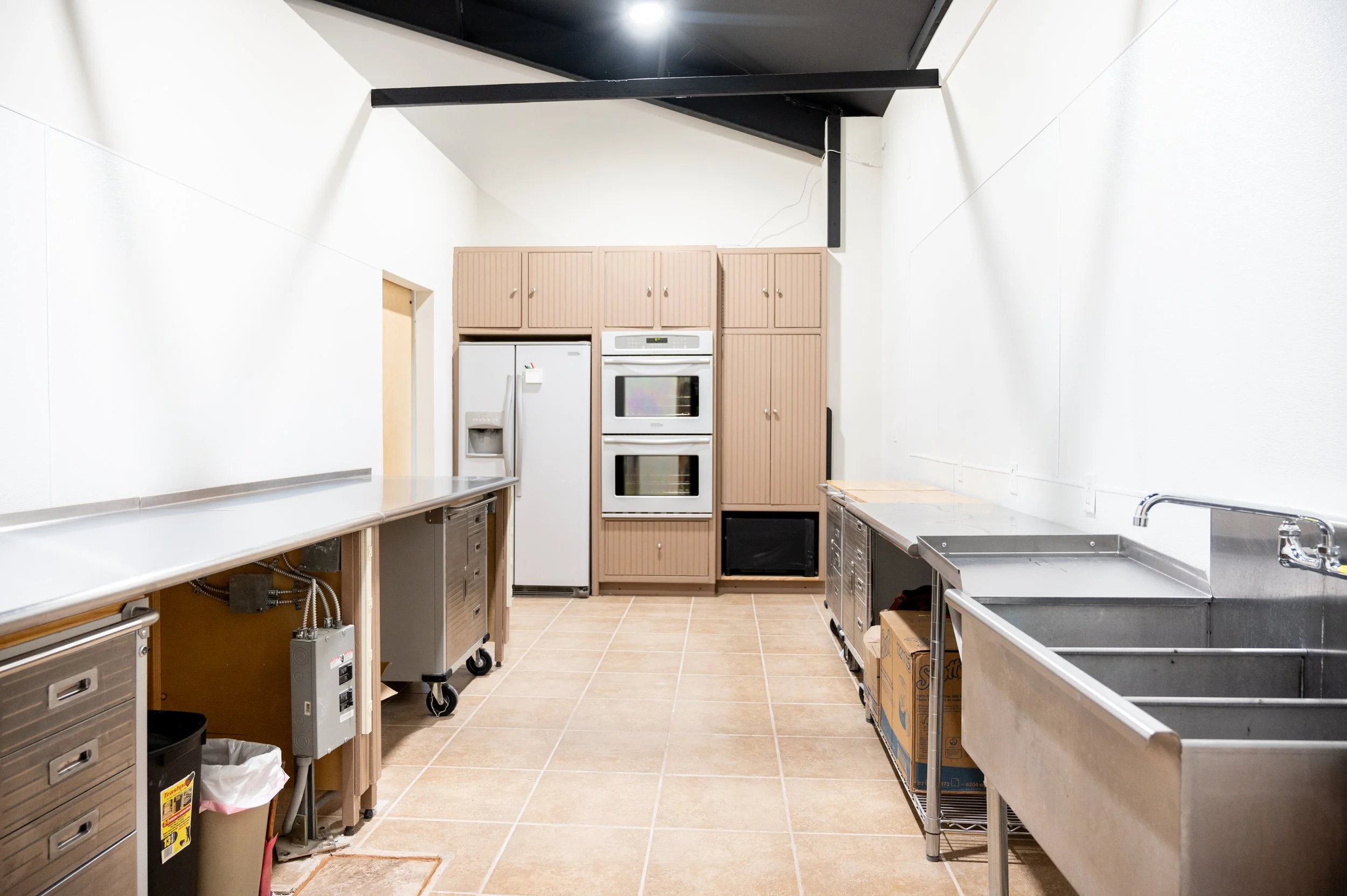 A commercial kitchen with stainless steel countertops, a double sink, and beige cabinets at the back, with a white refrigerator and wall ovens.