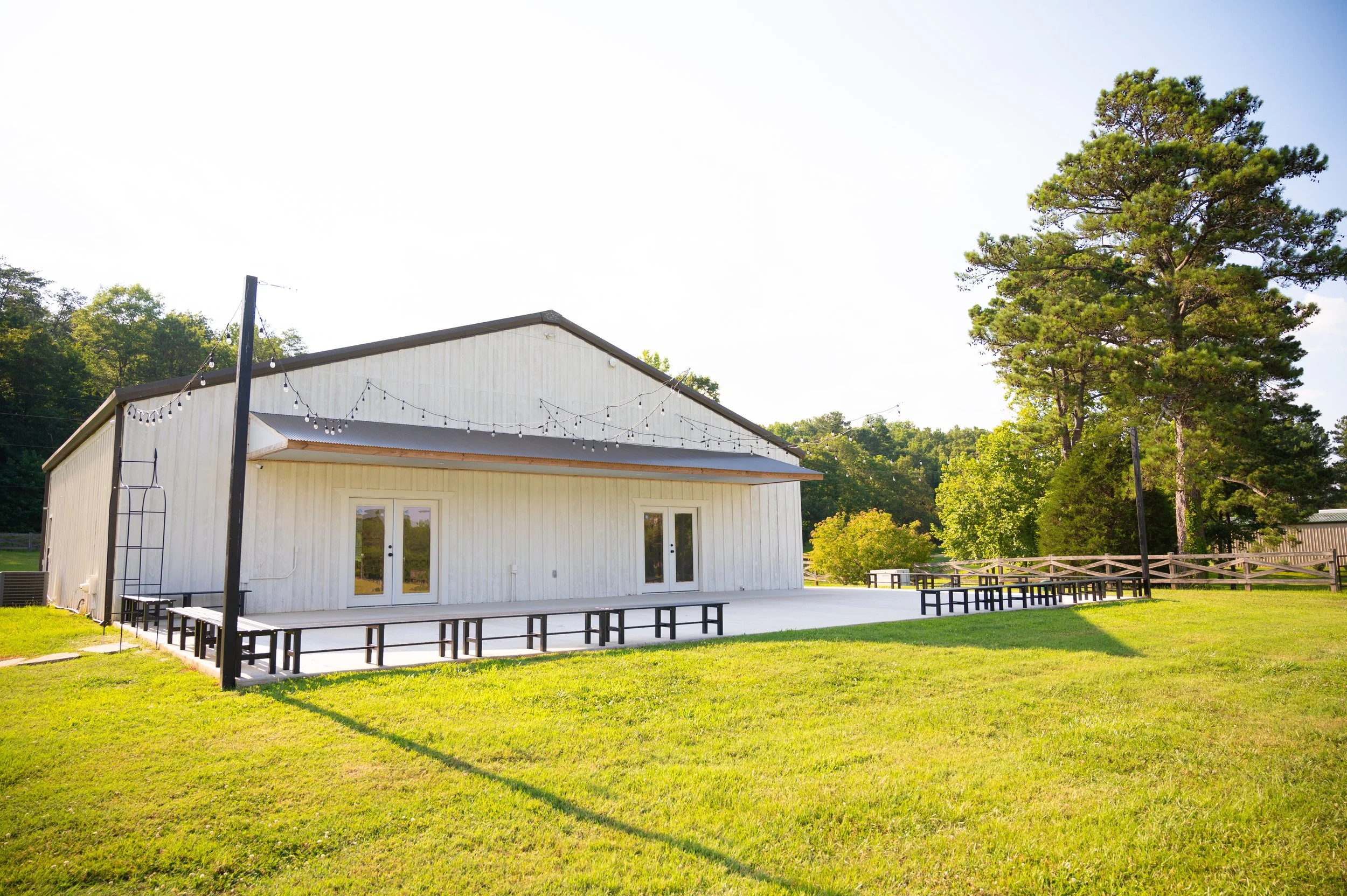 White building with string lights and black benches on a patio, surrounded by green grass and large trees.