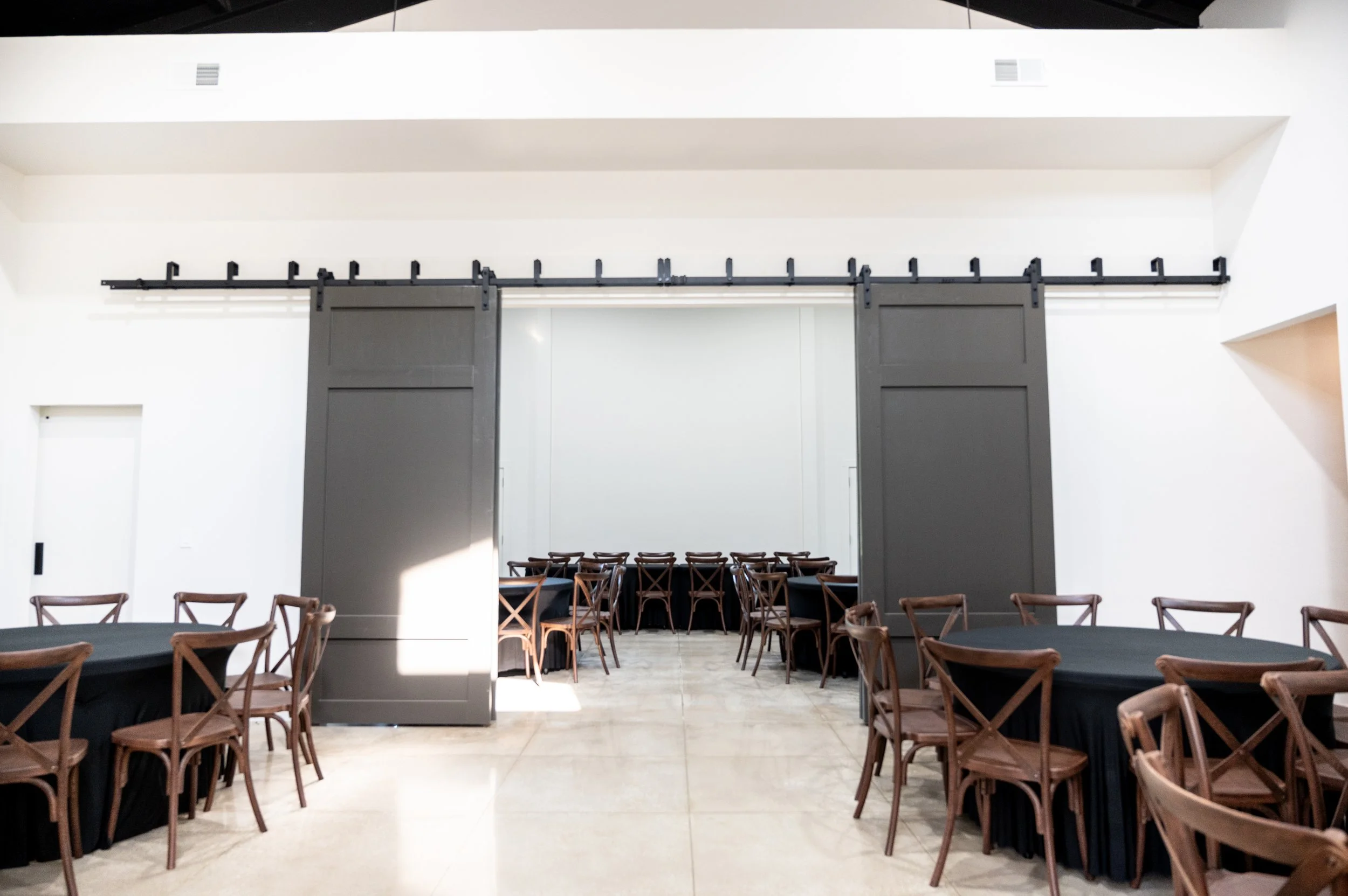 Modern banquet hall with round tables covered in black tablecloths, surrounded by wooden chairs, and large sliding barn doors in the background.