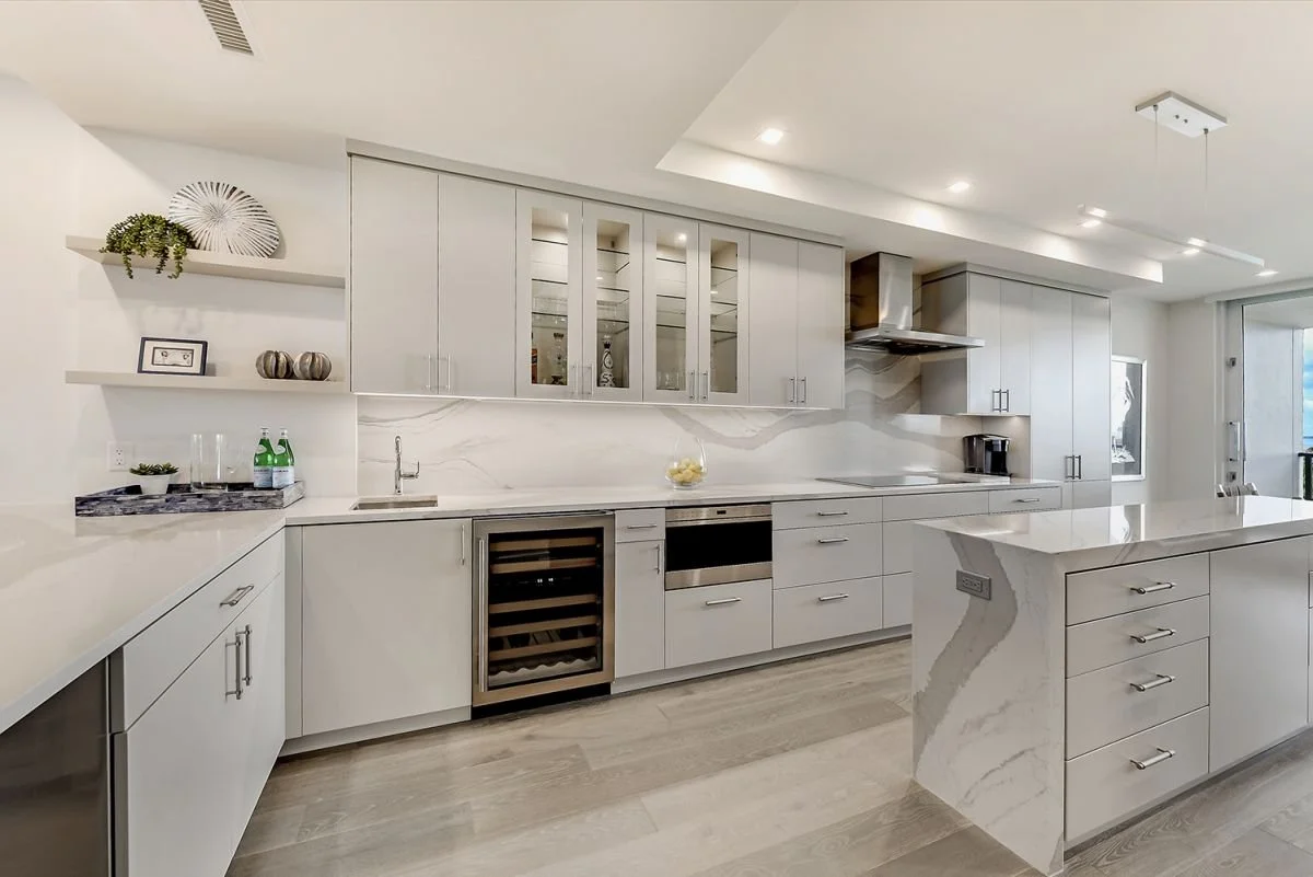 Modern white kitchen with marble countertops, upper cabinets with glass inserts, wine cooler, sink, stovetop, and bright lighting.