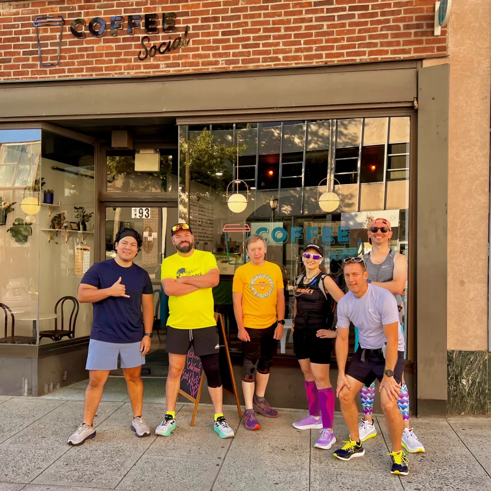 Group of six people standing outside a coffee shop called Coffee Social, smiling at the camera, some wearing athletic clothing.