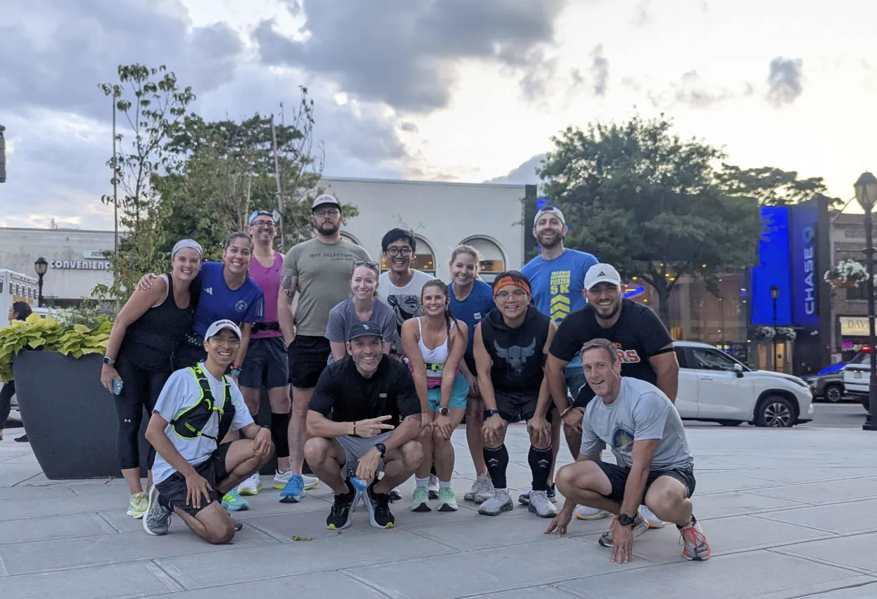 Group of 14 runners in athletic clothing and gear, posing together outdoors on city sidewalk with commercial buildings, trees, and cloudy sky in the background.