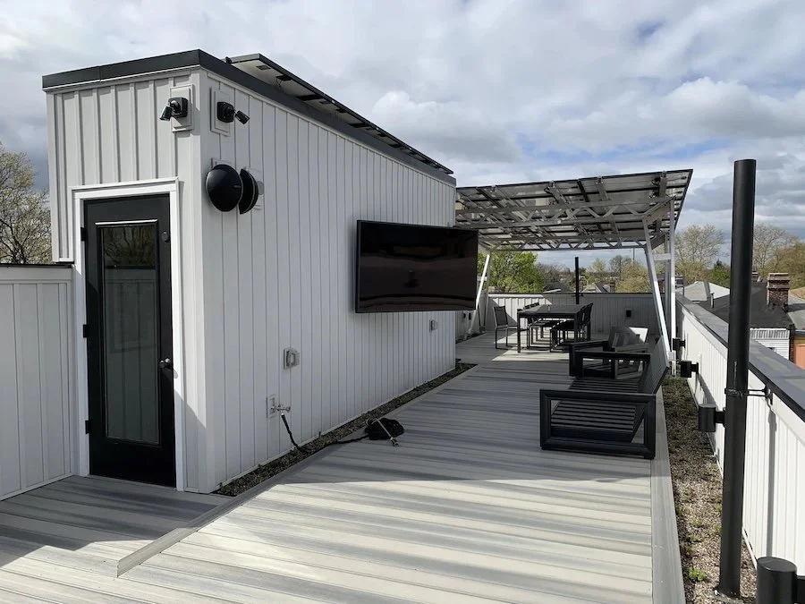 Rooftop deck with outdoor TV mounted on siding, security cameras, speakers, pergola with seating, and skyline view.