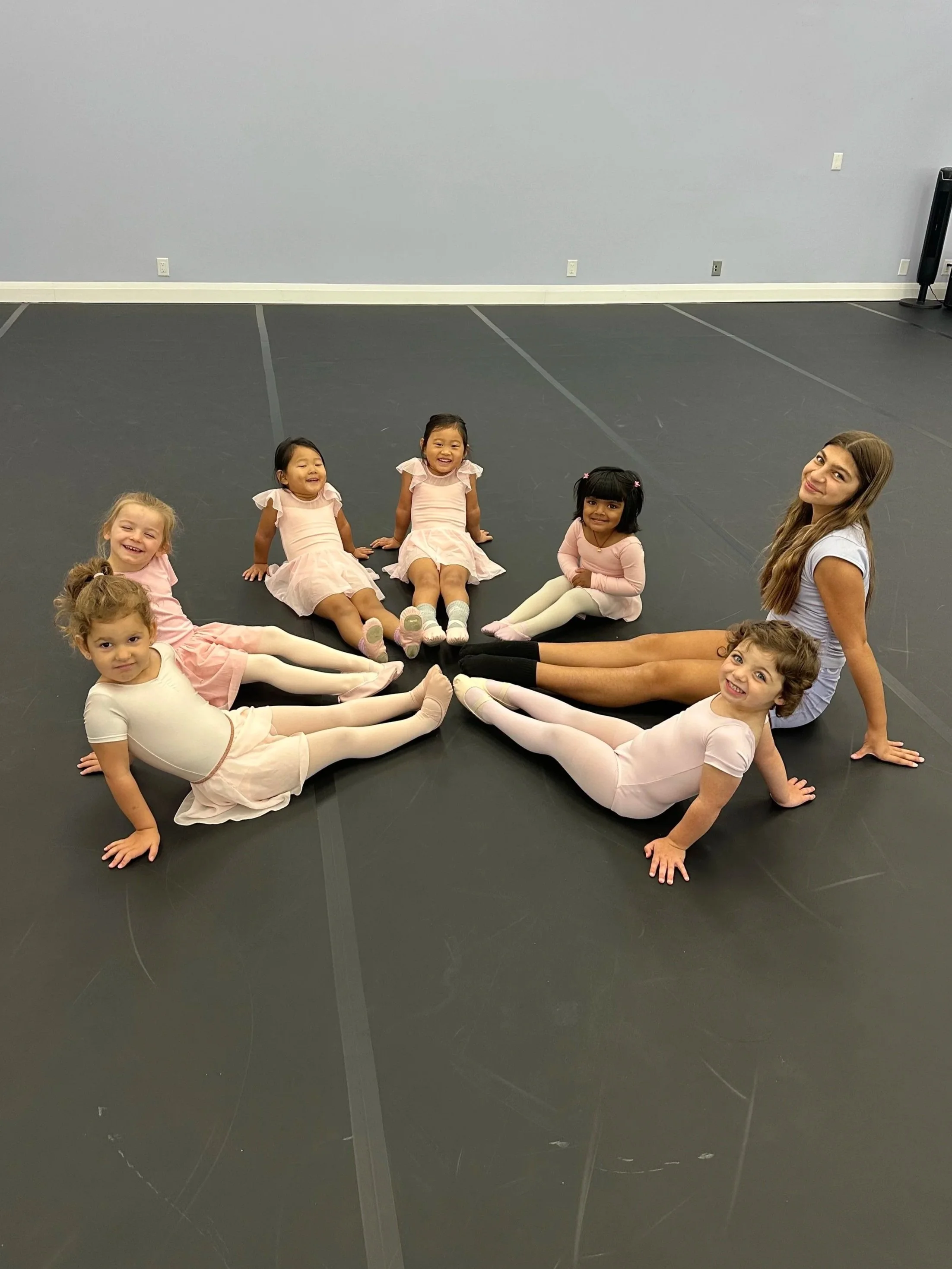 Child in pink tutu and socks on stage with arms outstretched, facing a lit theater with ornate ceiling and chandelier.