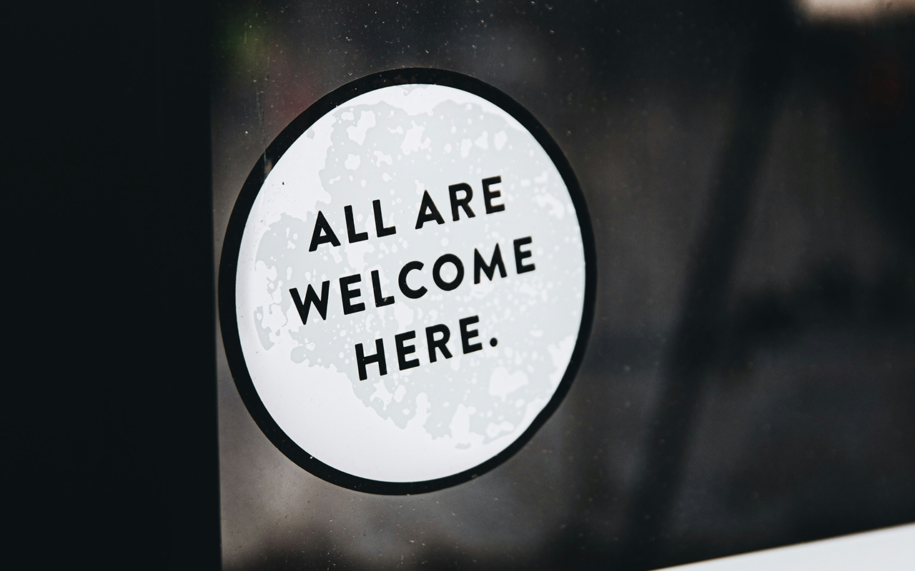 Sticker that reads ‘All are welcome here’ on a dark glass door