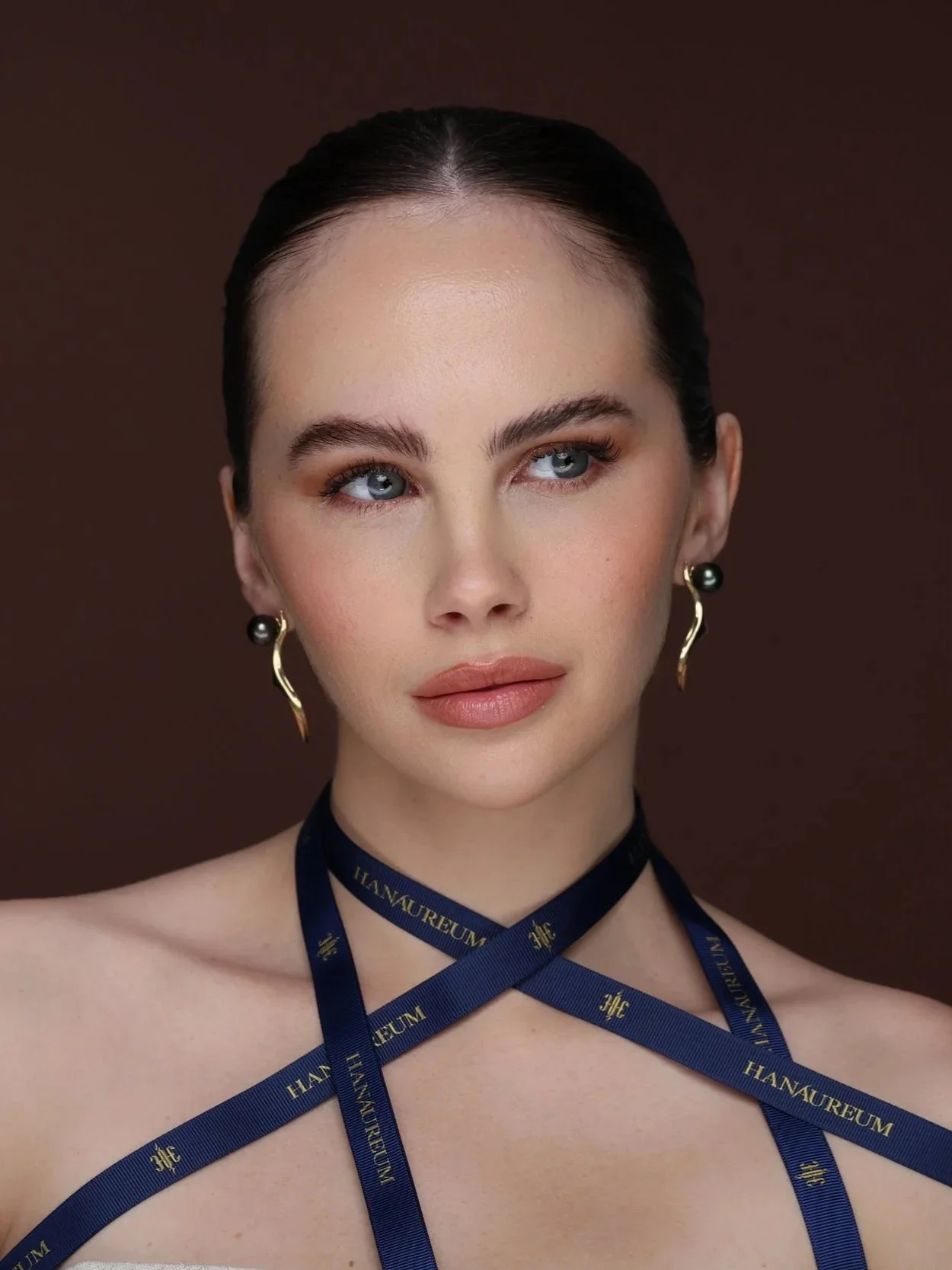 Front-facing portrait of a model wearing HANAUREUM Sylven earrings in 14K gold with Tahitian pearls and sculptural antler-inspired forms, styled with navy ribbon.