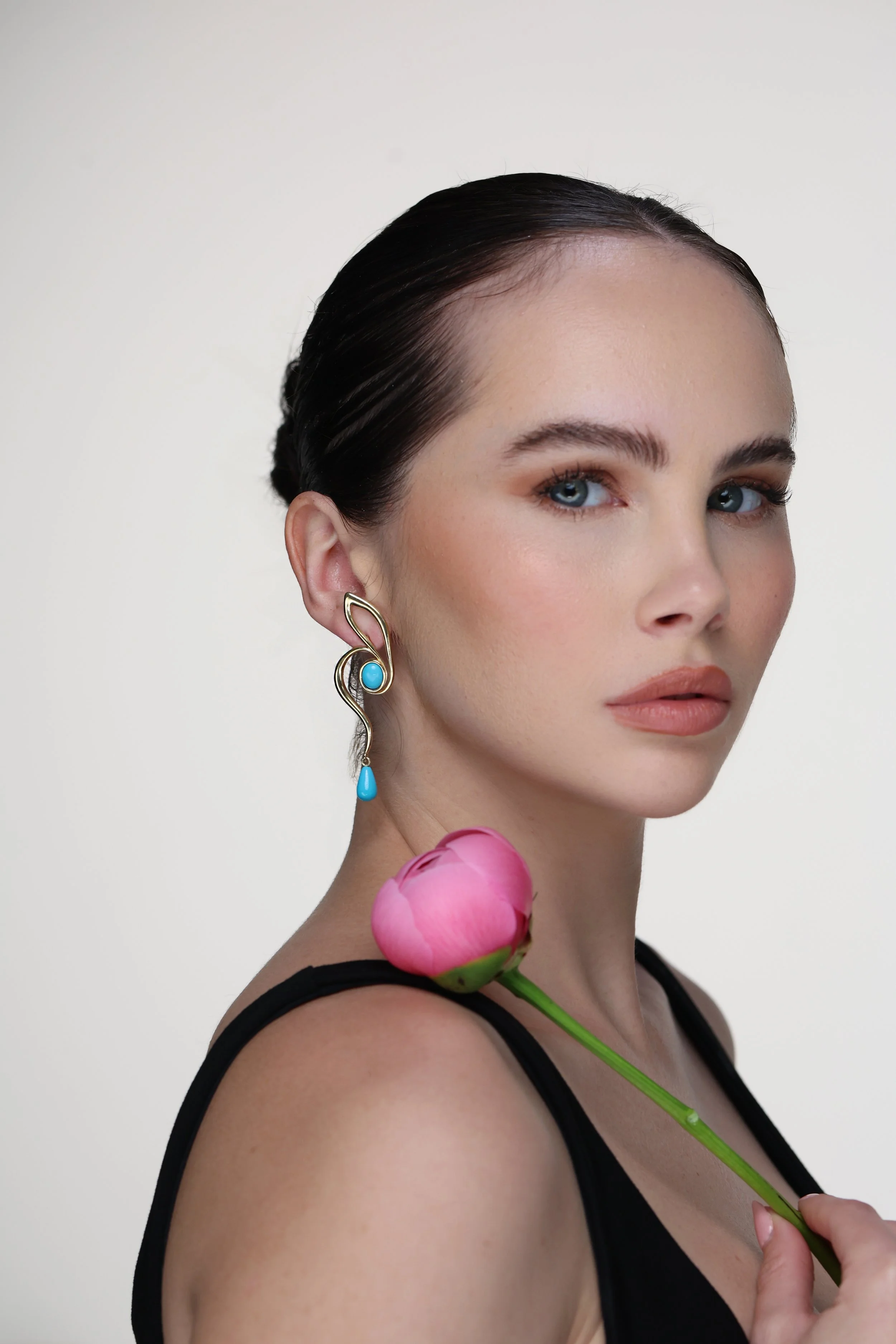 Portrait of a model holding a pink peony flower and wearing the Lumi 14K gold earring with oval and teardrop turquoise stones