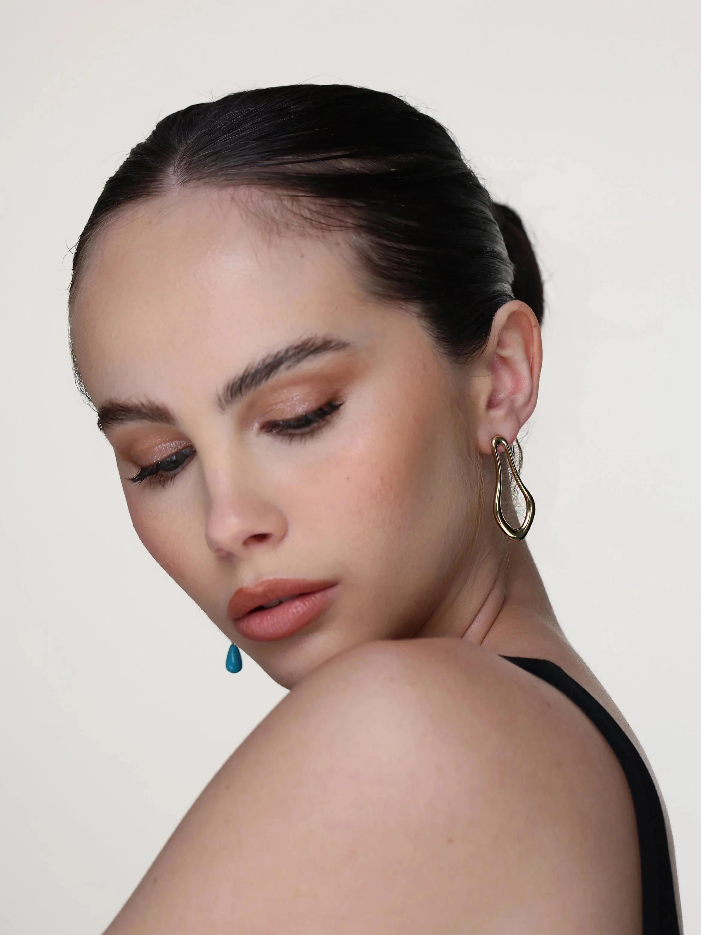 Close-up of a Model with dark hair wearing HANAUREUM LUMI sculptural earrings in 14K gold with a turquoise accent against a plain white background.
