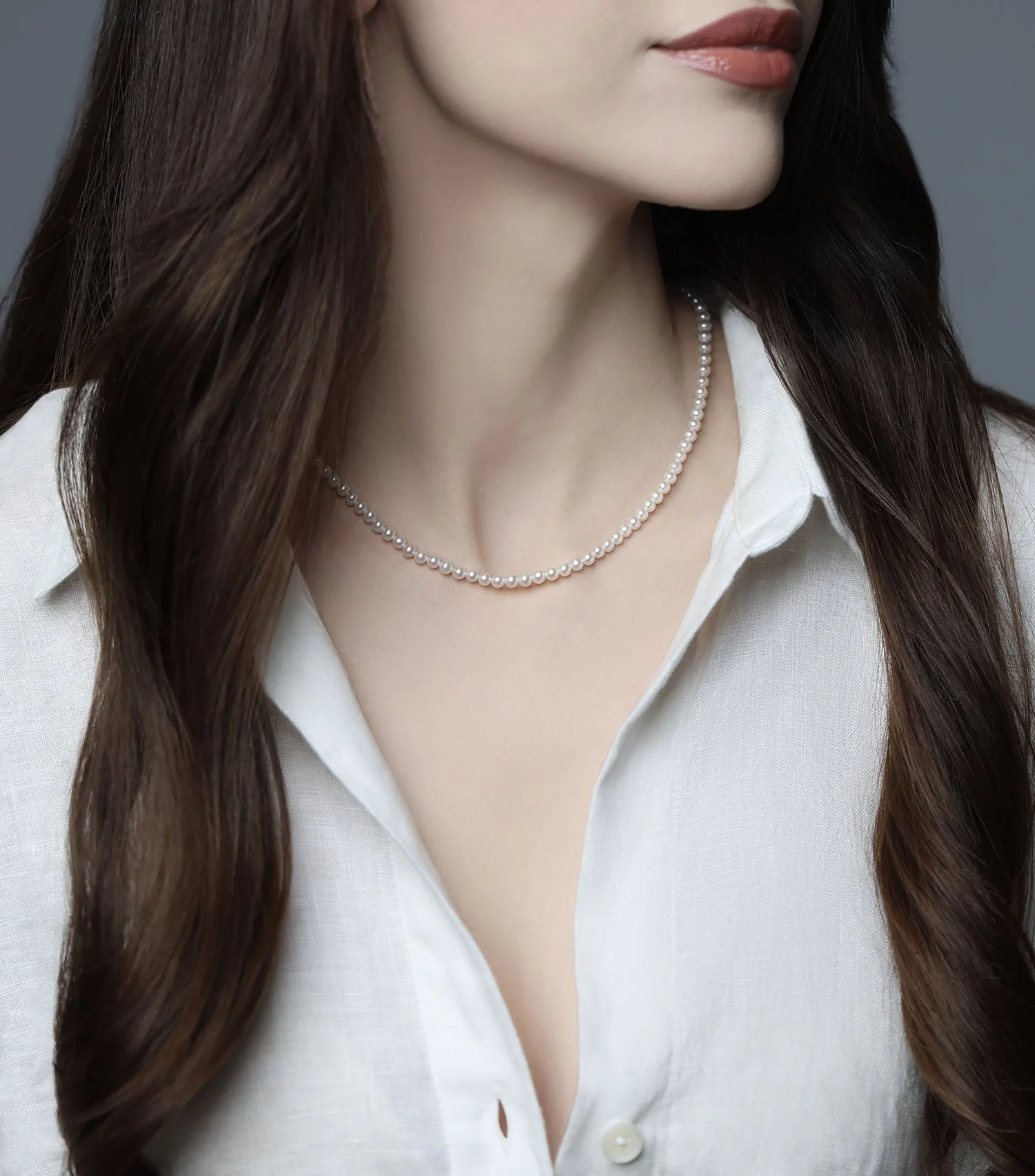 Model with long wavy dark brown hair and a white button-down blouse wears the Élan 3 mm cultured white pearl necklace at her collarbone against a soft gray background.