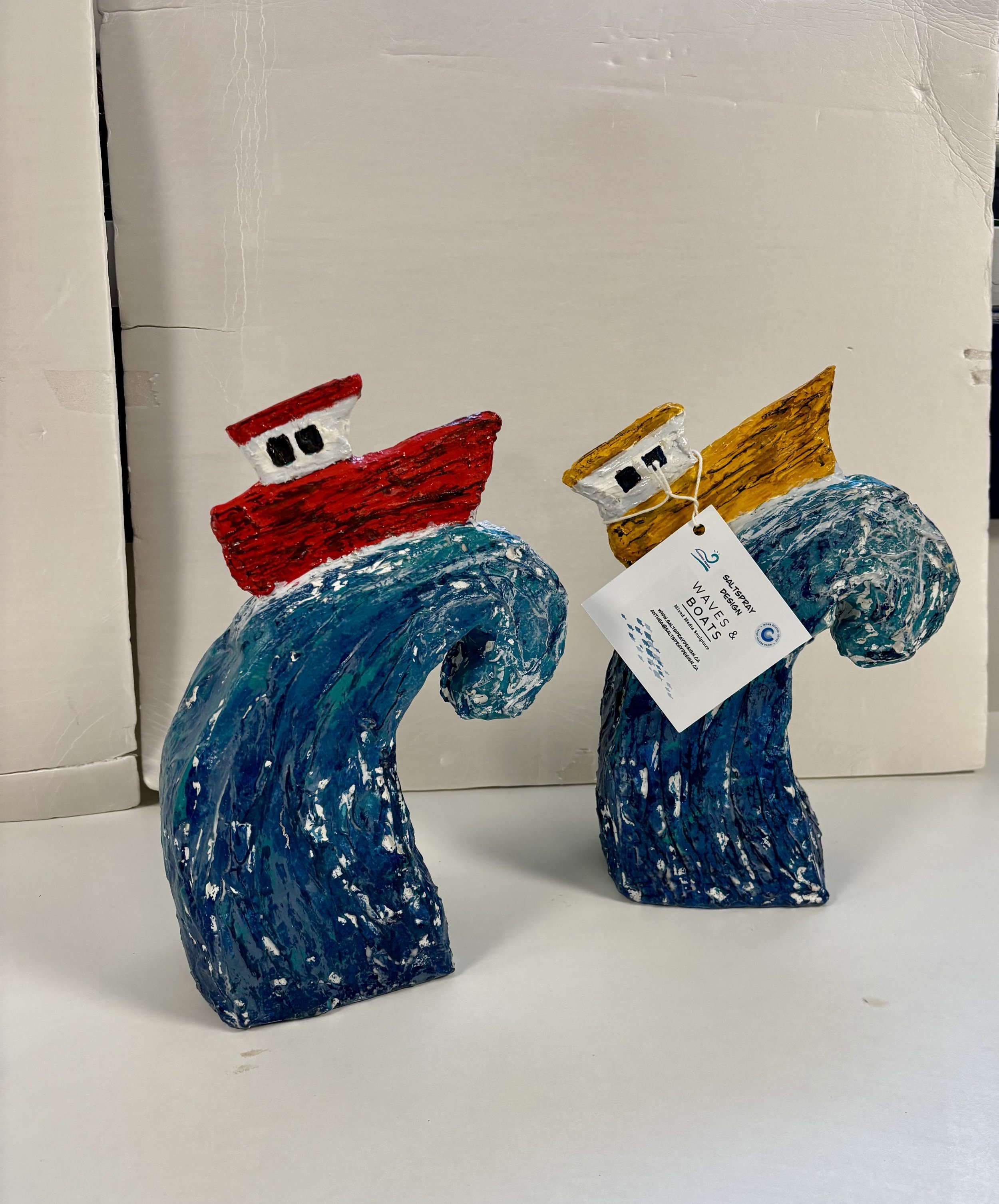 Anthea Taljaard - SaltSpray Design
Two colorful boat sculptures riding ocean waves, made of textured material with a label attached to one, displayed against a plain background.