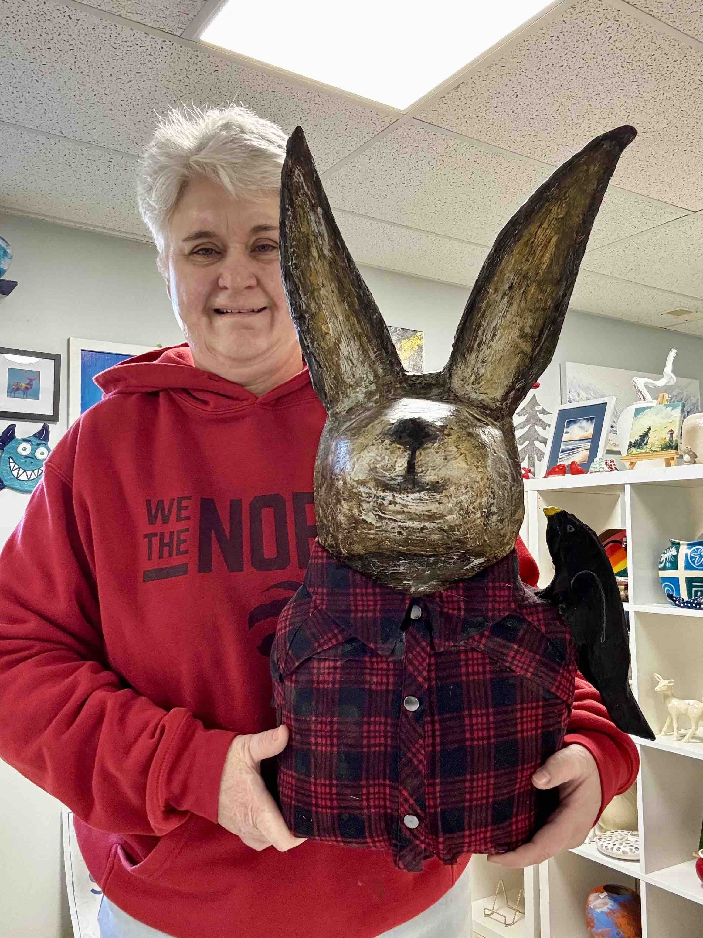 Anthea Taljaard - SaltSpray Design
Person holding large rabbit sculpture wearing a red plaid shirt, with a black bird perched on its shoulder, indoors with art and decorations in the background.