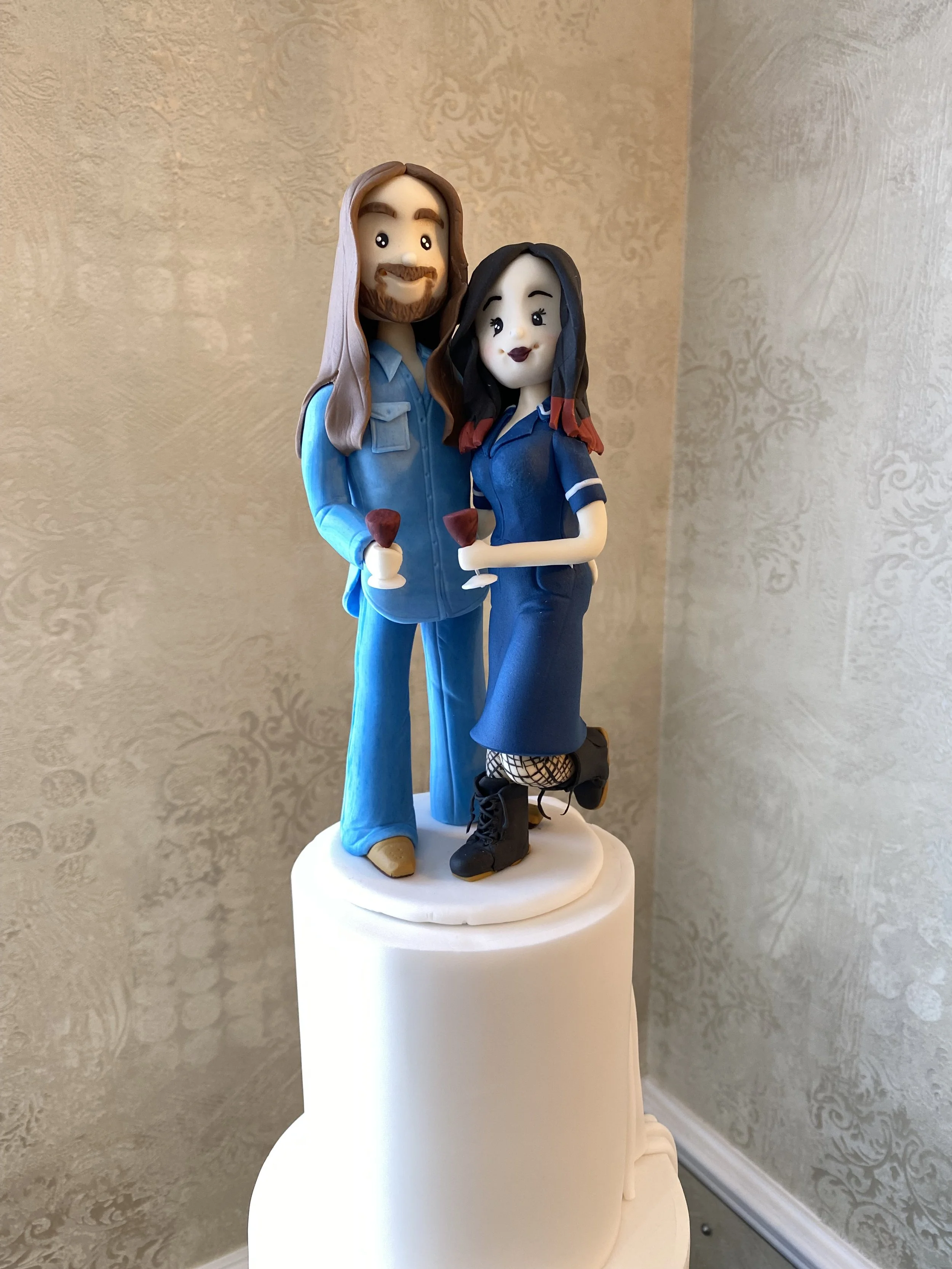 Alternative bride and groom cake figurine, groom with double denim and long hair, bride dressed as nurse with DrMarten boots. Both holding red wine glasses