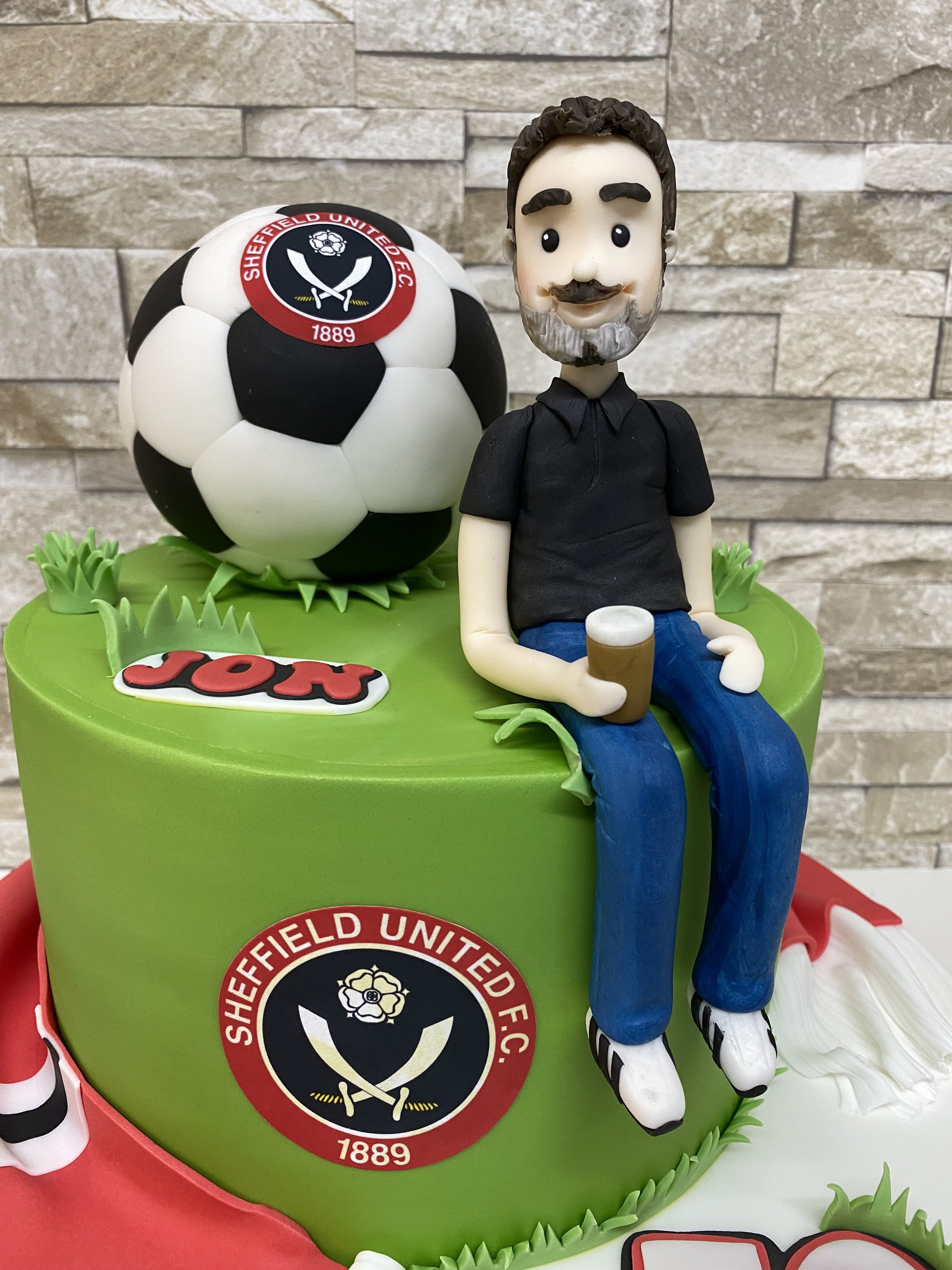 Male cake figurine seated on a green birthday cake holding a glass of beer, with Sheffield United football 