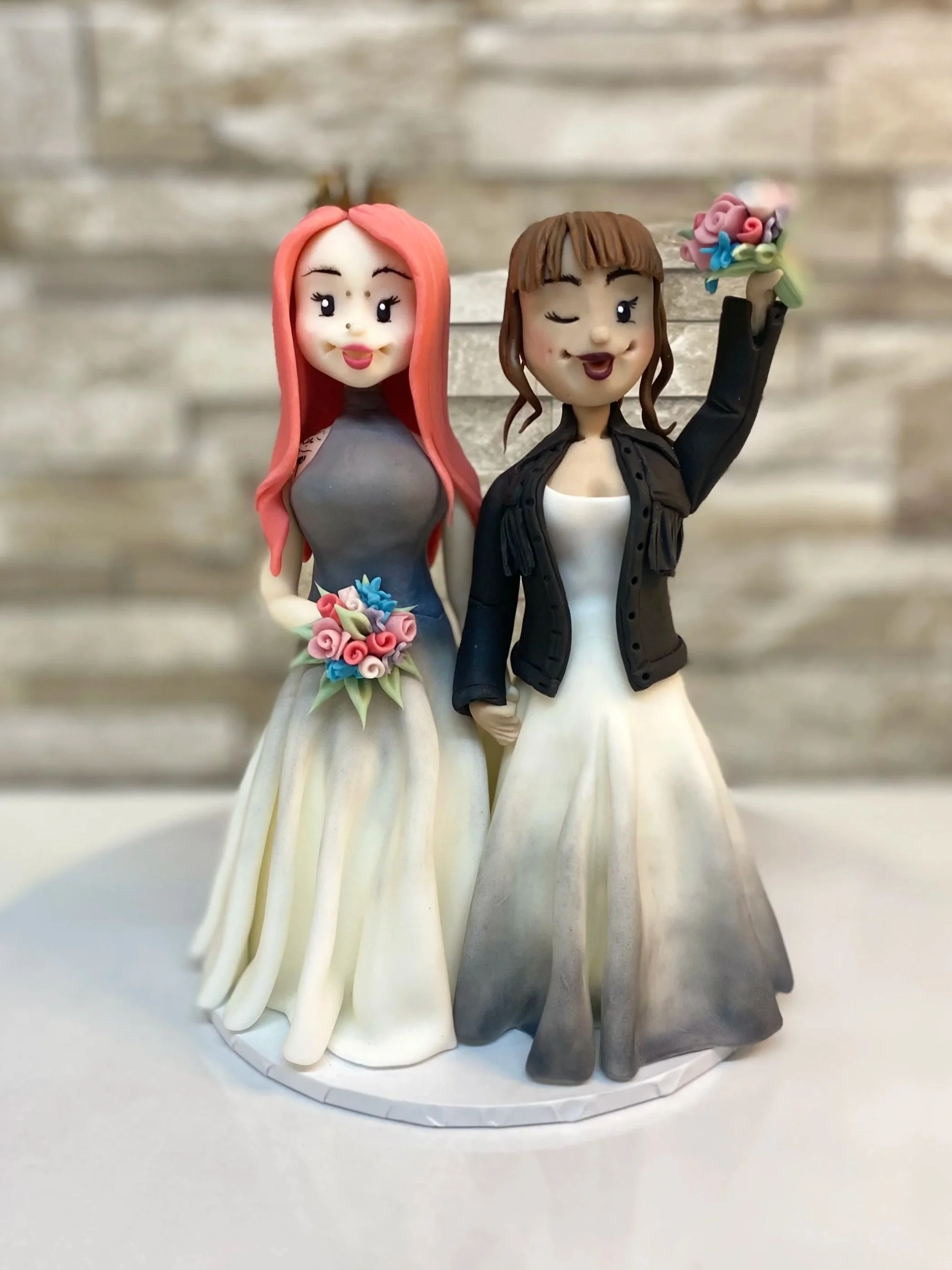 Bride and Bride cake topper both wearing Lucy Can't Dance couture dresses with leather jacket and facial piercings. One with arm in the air and one with pink hair
