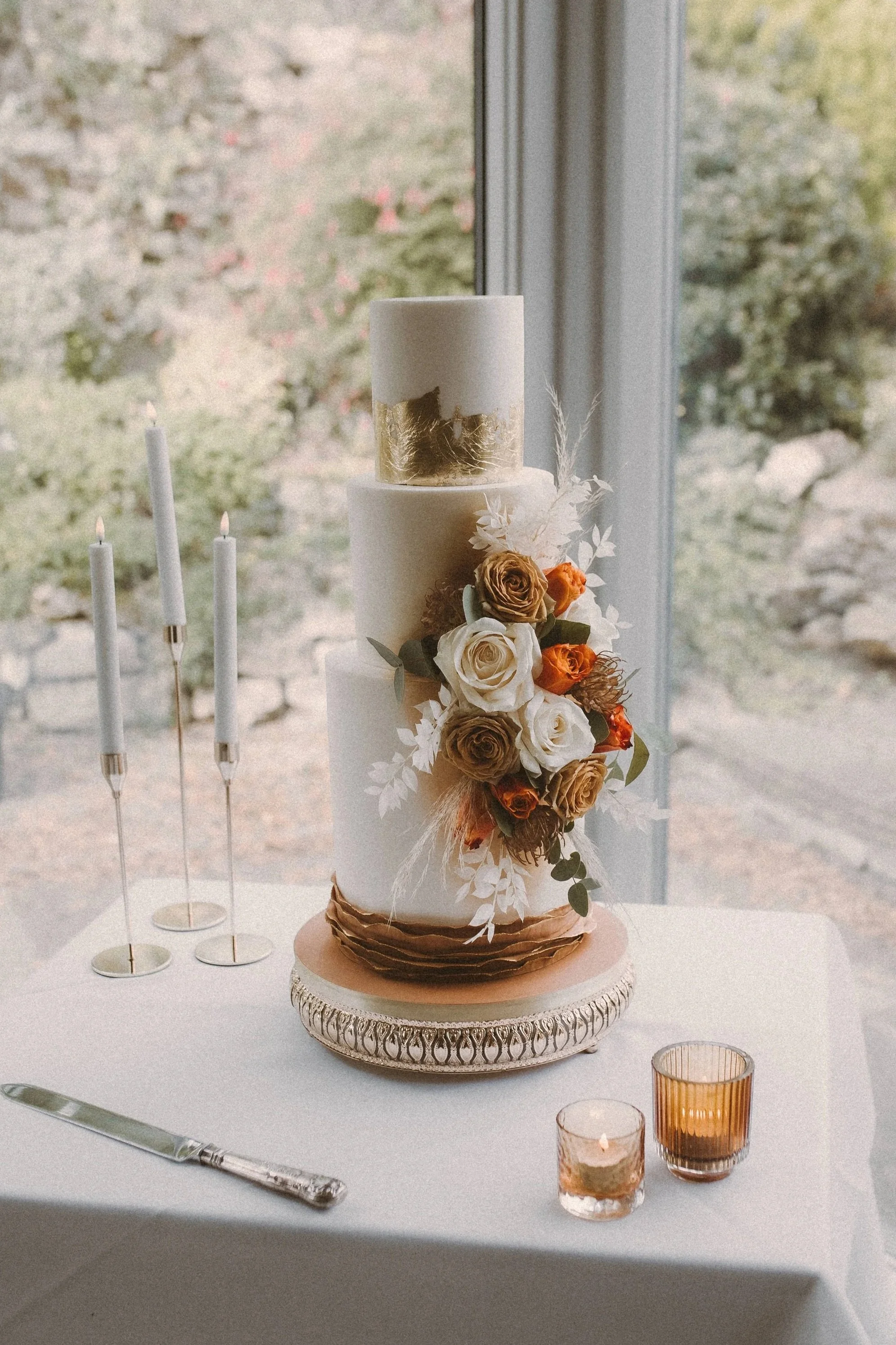 three tiered wedding cake with extra tall bottom tier. Autumnal colour palette and gold leaf highlights. Statement flower decoration to the side. Placed on a gold cake stand on a table with tall candle sticks and tea lights in front of a window at We