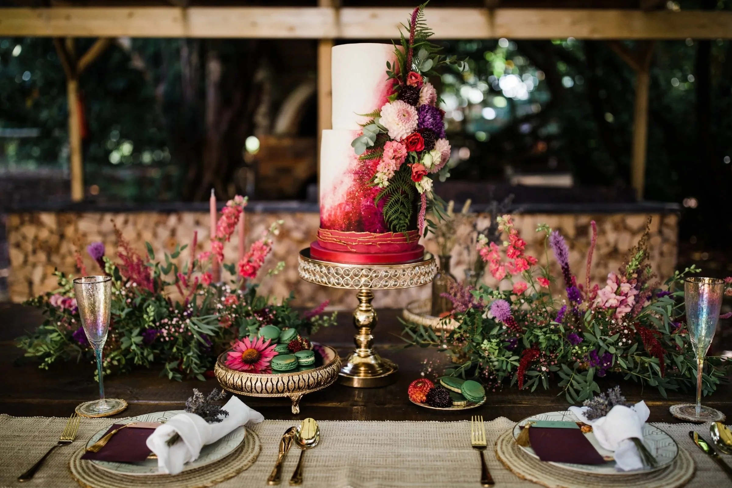 Decorative wedding cake with floral arrangements on a table surrounded by floral centerpieces, macarons, champagne flutes, plates with napkins, and cutlery.