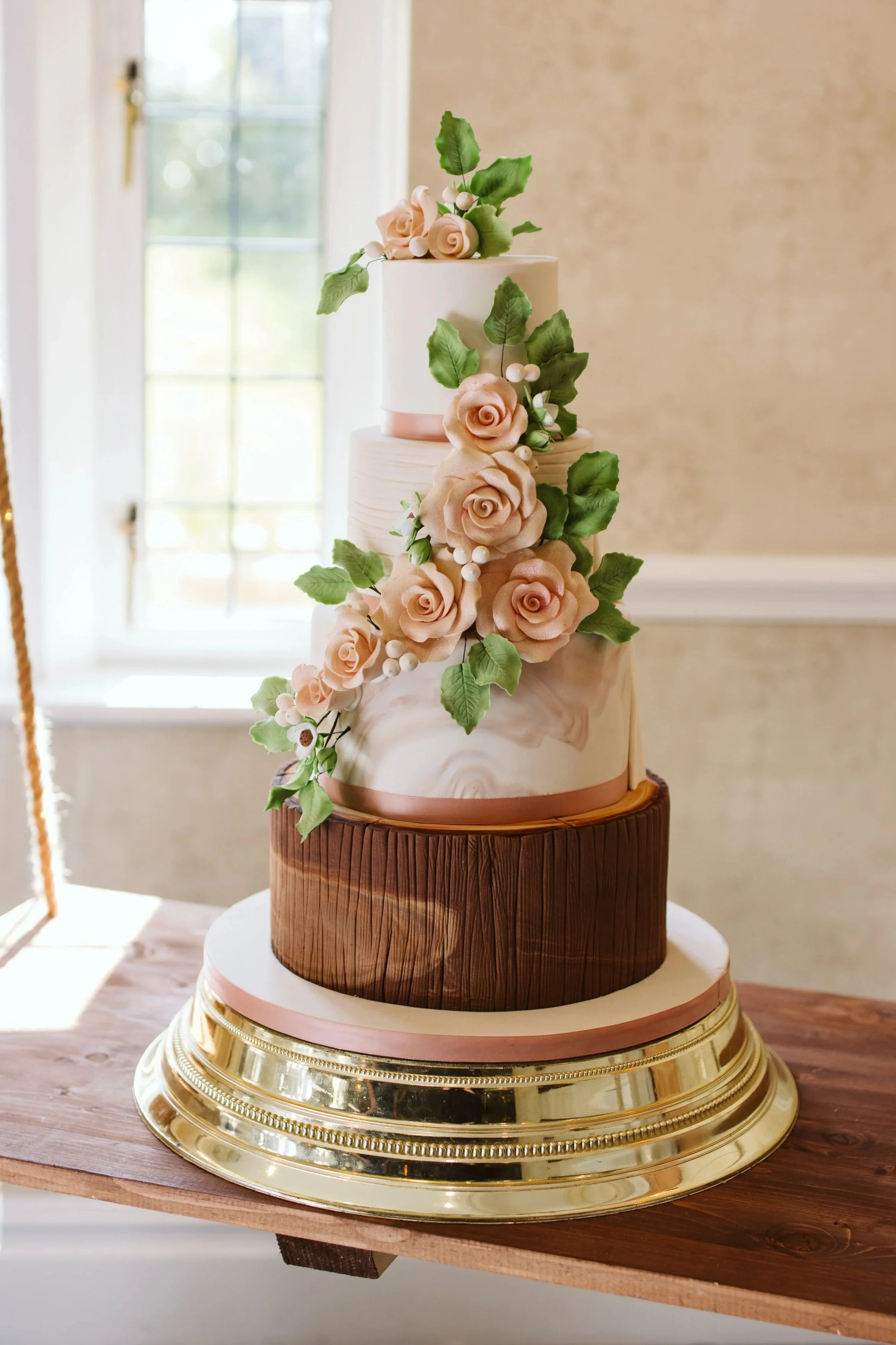 four tier wedding cake with  marble tier and log bottom tier, cascade of blush pink sugar roses and leaves. Placed on a gold cake stand on a cake swing in front of the window at Rogerthorpe Manor Hotel