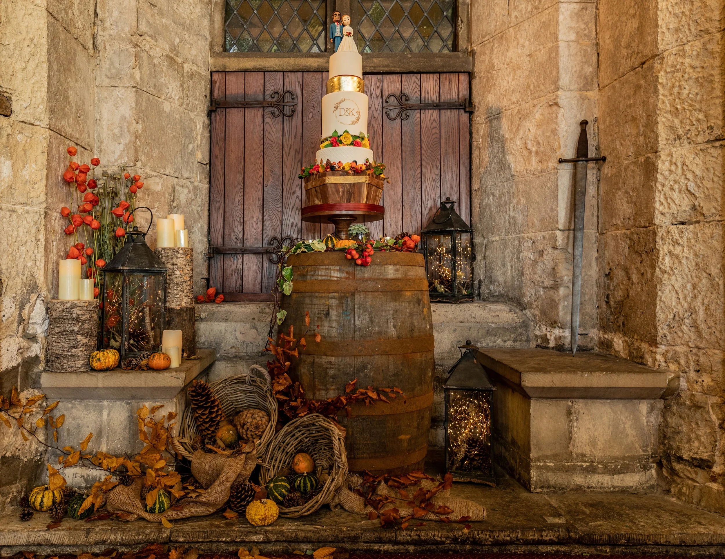 Tall autumn wedding cake displayed on a wooden barrel in a rustic stone setting, decorated with gold detailing and seasonal flowers, surrounded by lanterns, candles, pumpkins, fallen leaves, and woven baskets for a warm harvest-themed backdrop.