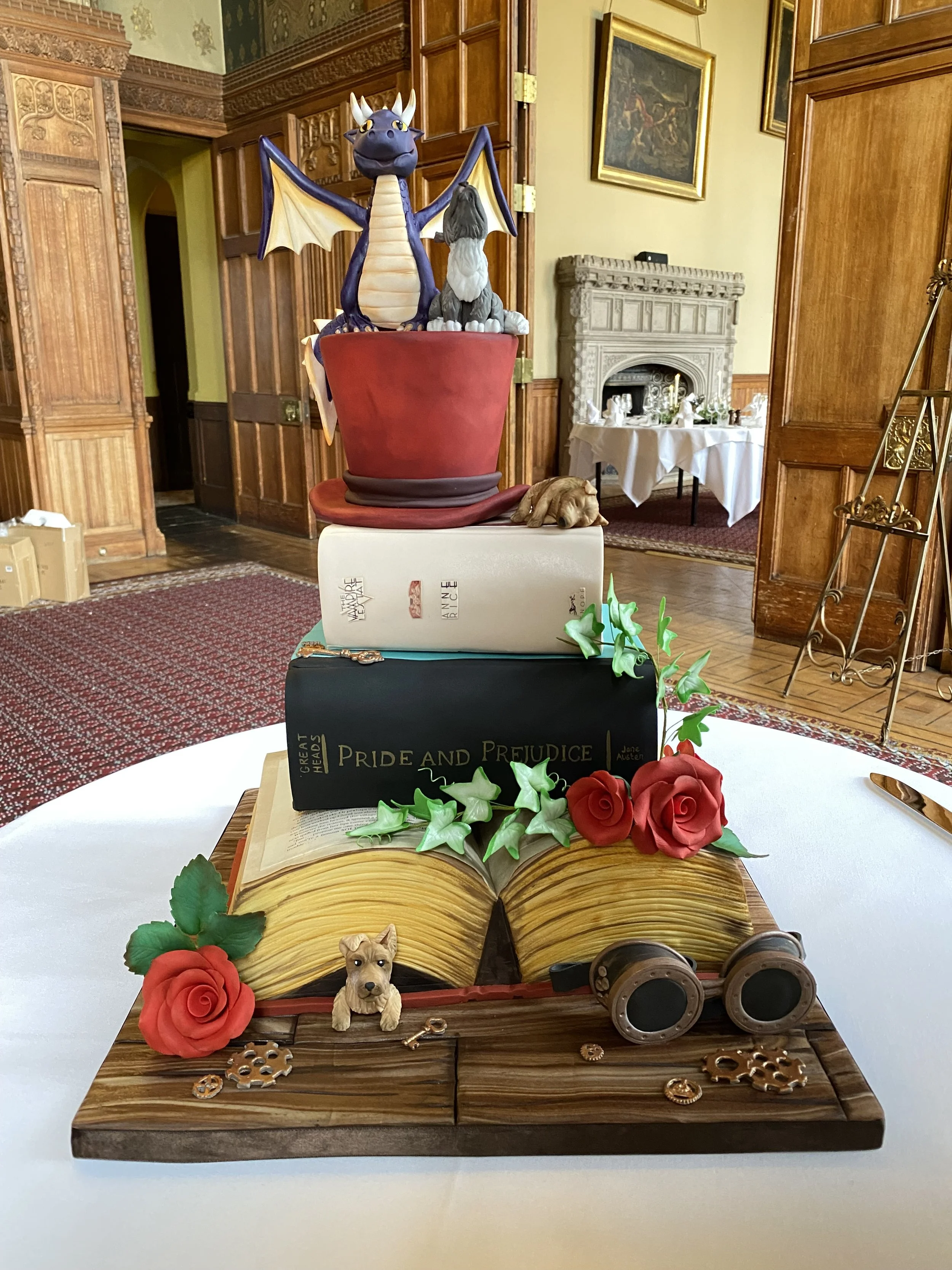 four tier alternative wedding cake, featuring an open book, two closed books and a red top hat. Steampunk paraphernalia, dogs and red roses decorate. Purple dragon and wolf cake topper set up in a gothic wood panelled room at Carlton Towers 