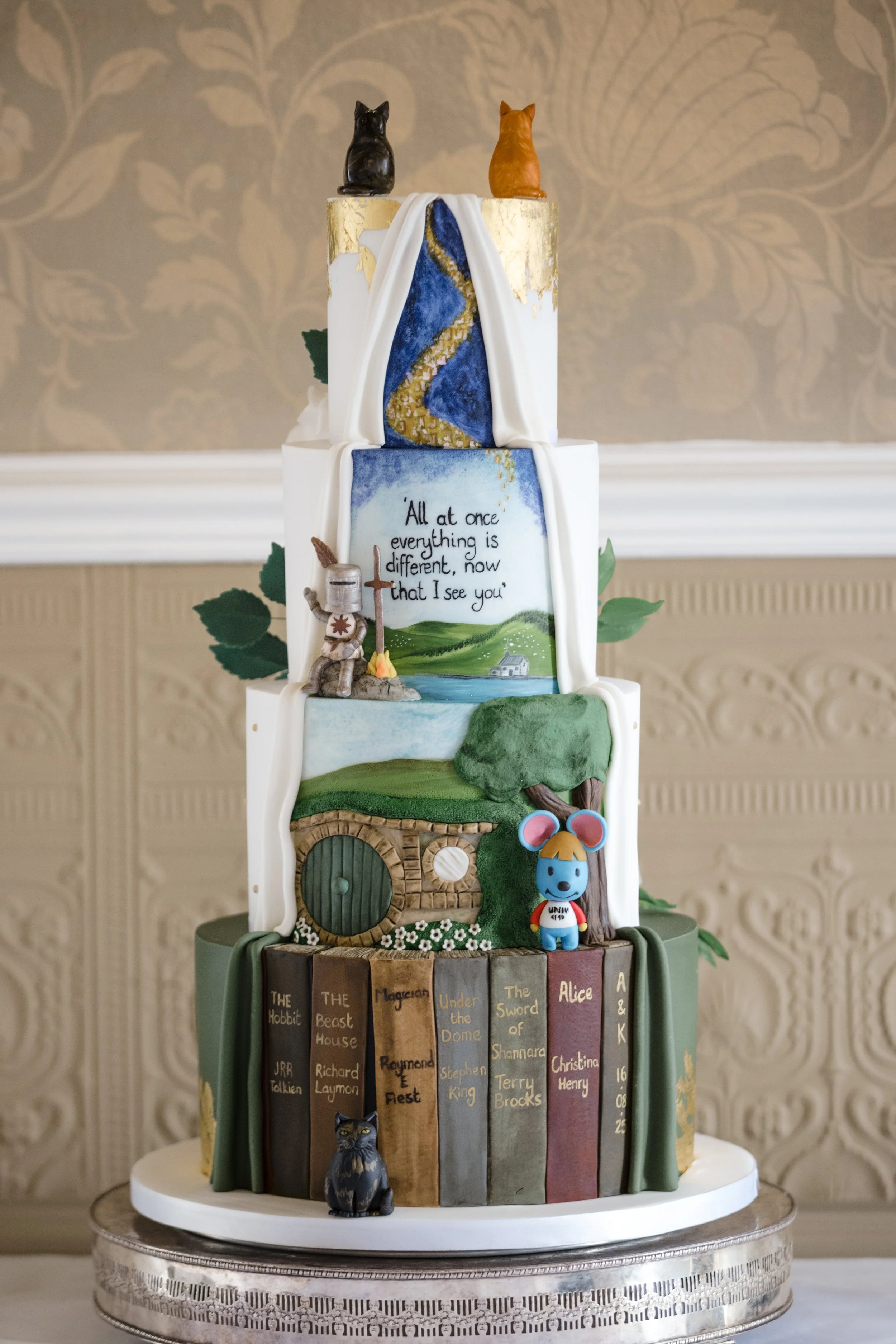Back side of a four tier wedding cake, featuring a library of books, The Hobbit house, and quote from Disney movie Tangled