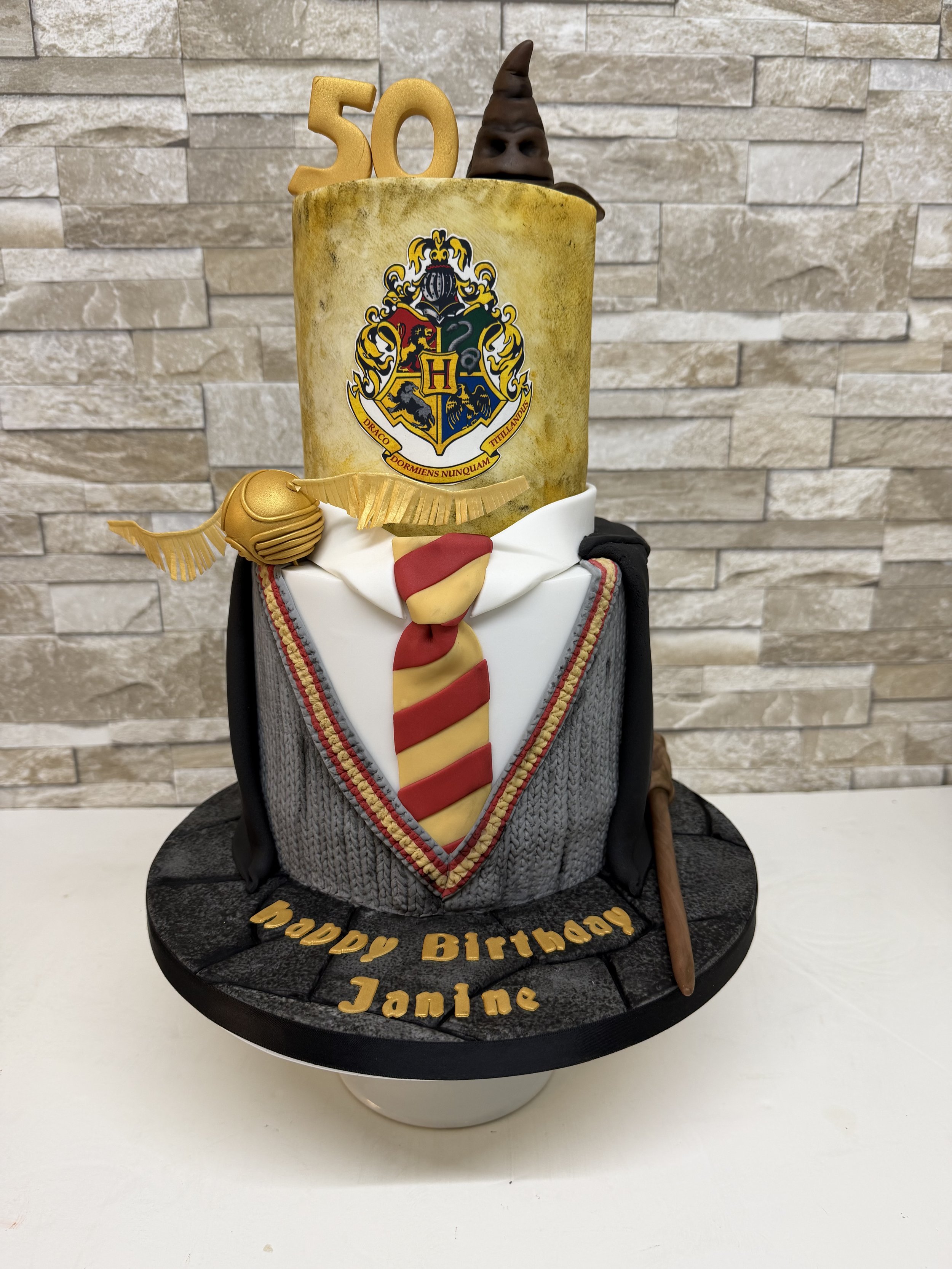 Two tier birthday cake with a Harry Potter theme. Gryffindor uniform bottom tier with sorting hat, wand and golden snitch