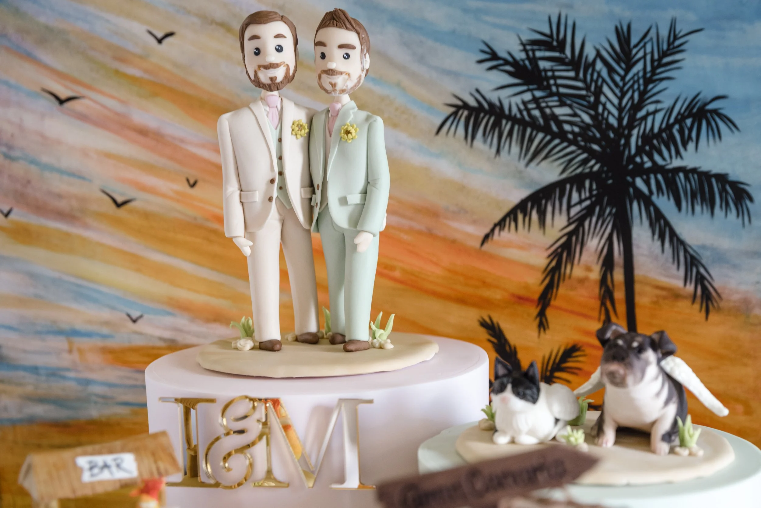 Male and male groom wedding cake topper in front of a  sugar painted sunset scene with cat and dog figurines to the side
