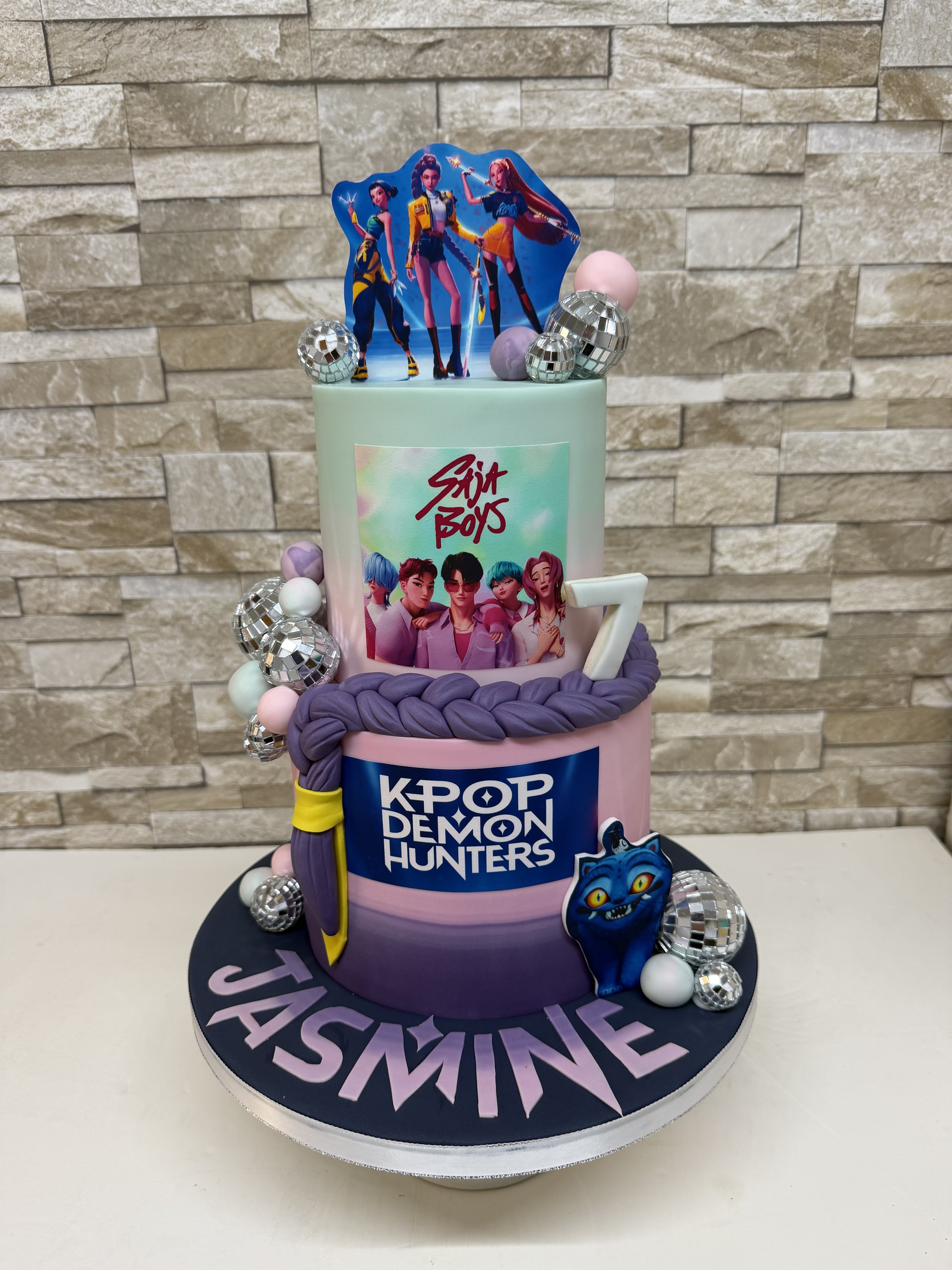 Two tier birthday cake with KPop Demon Hunters theme. Purple and blue coloured tiers with purple platt, glitter balls and character images