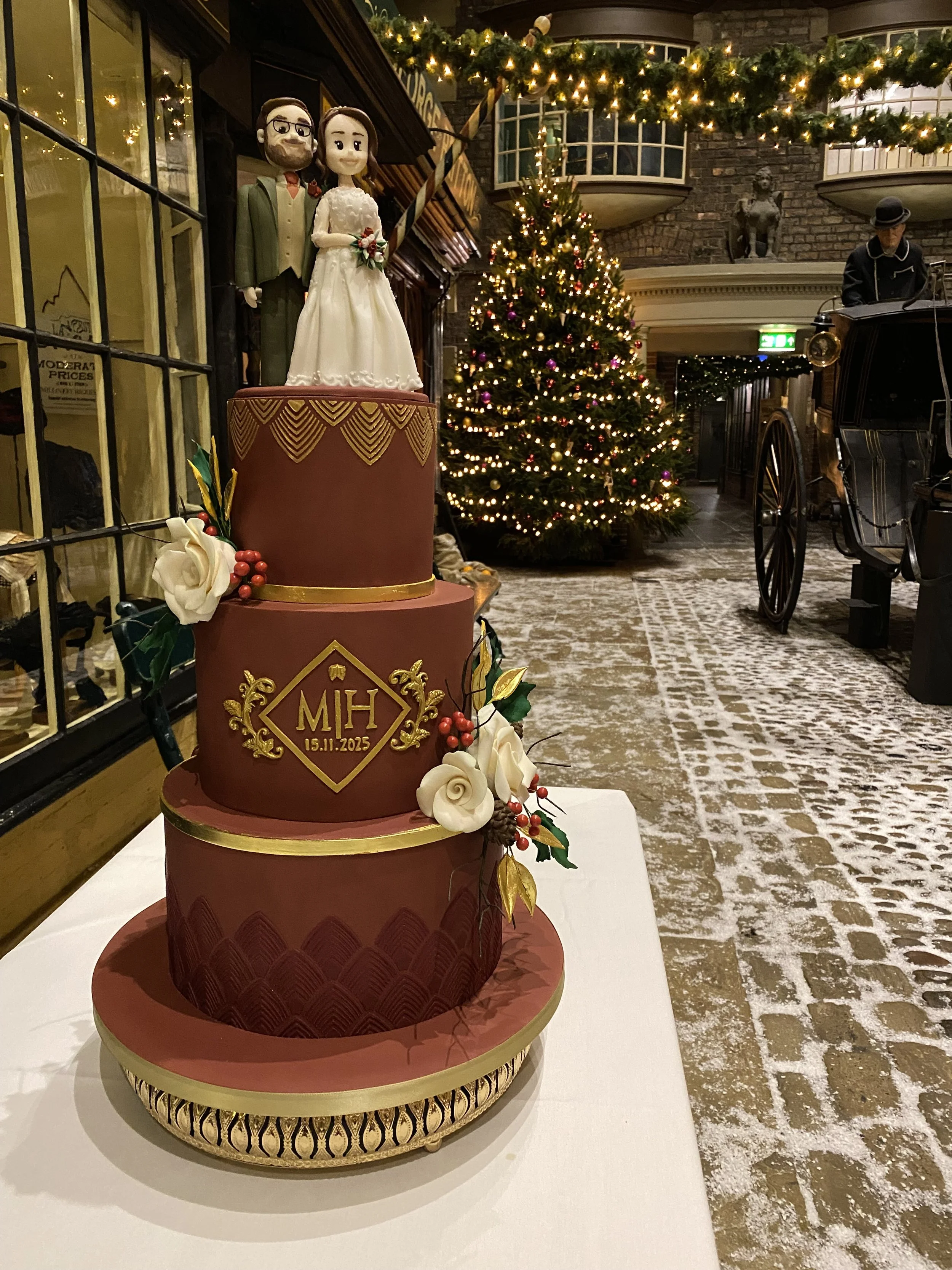 Burgundy three tier wedding cake with gold monogram and winter sugar flowers, bride and groom cake topper. Sat on a table in the Victorian street at York Castle Museum during Christmas time  