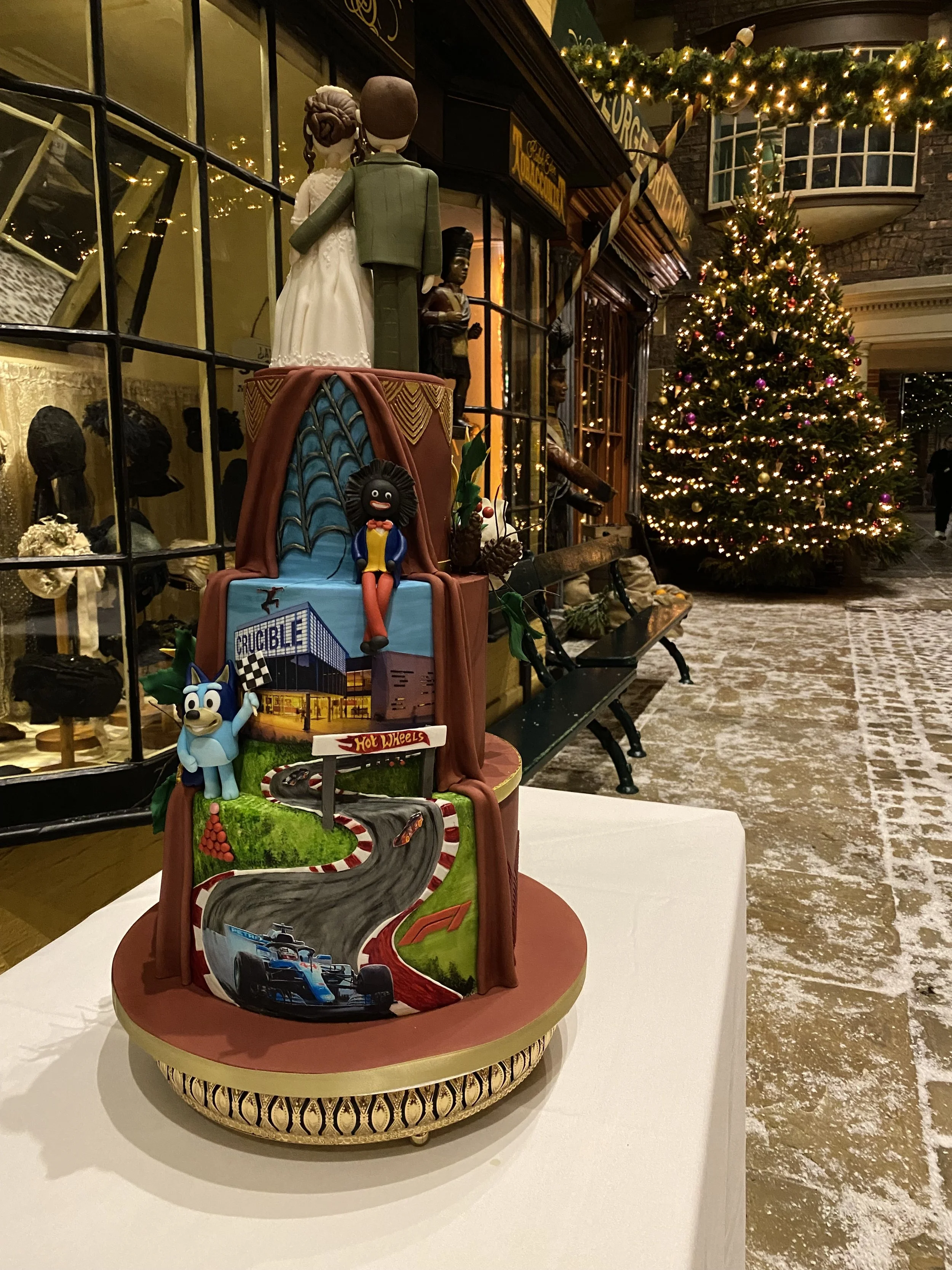 Back reveal side of a three tier wedding cake with formula one, snooker, and Bluey theme. Sat on a table in the Victorian street at York Castle Museum during Christmas time