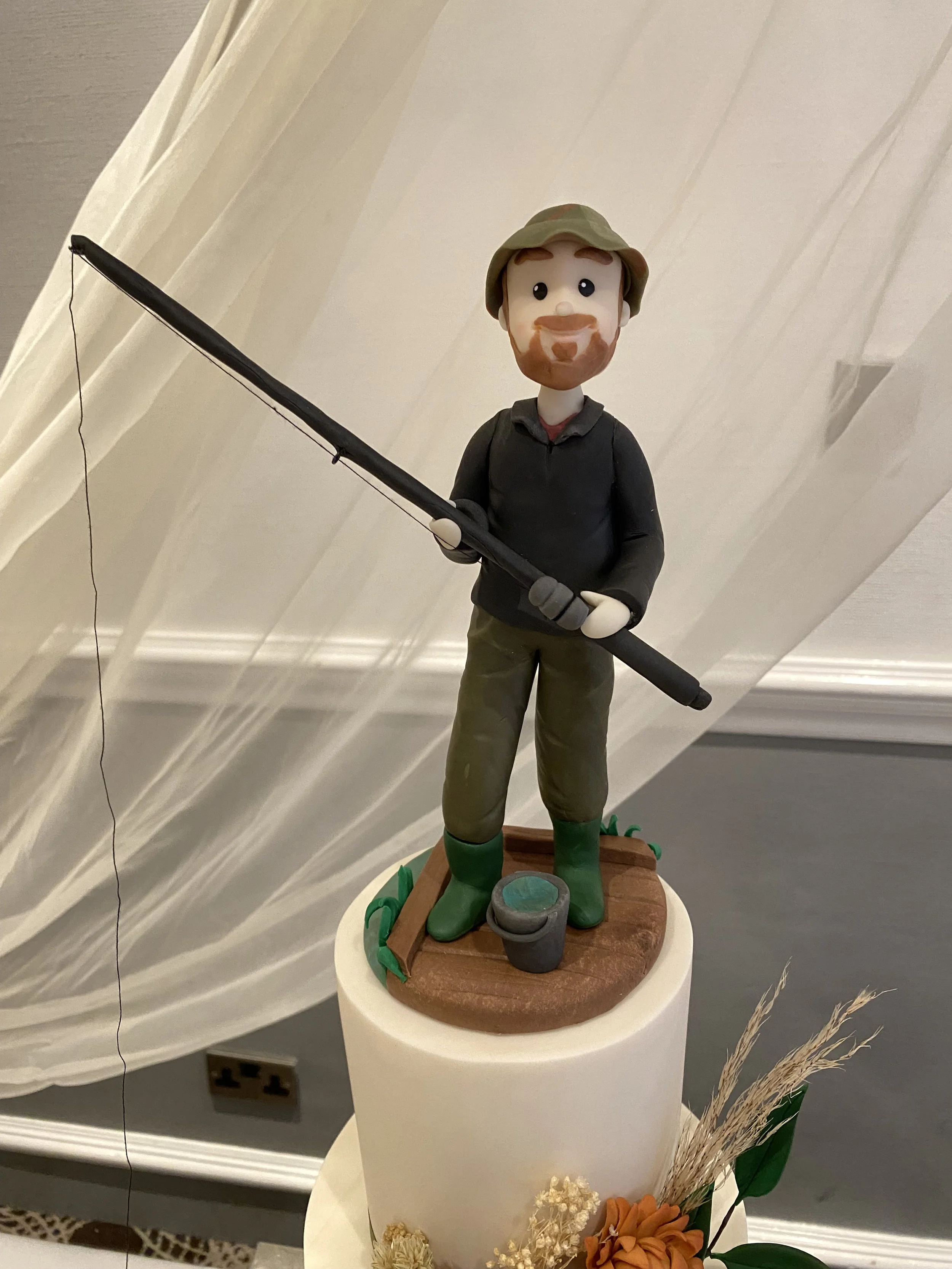 Single male cake figurine dressed in fishing clothes and fishing rod situated at the top of a wedding cake