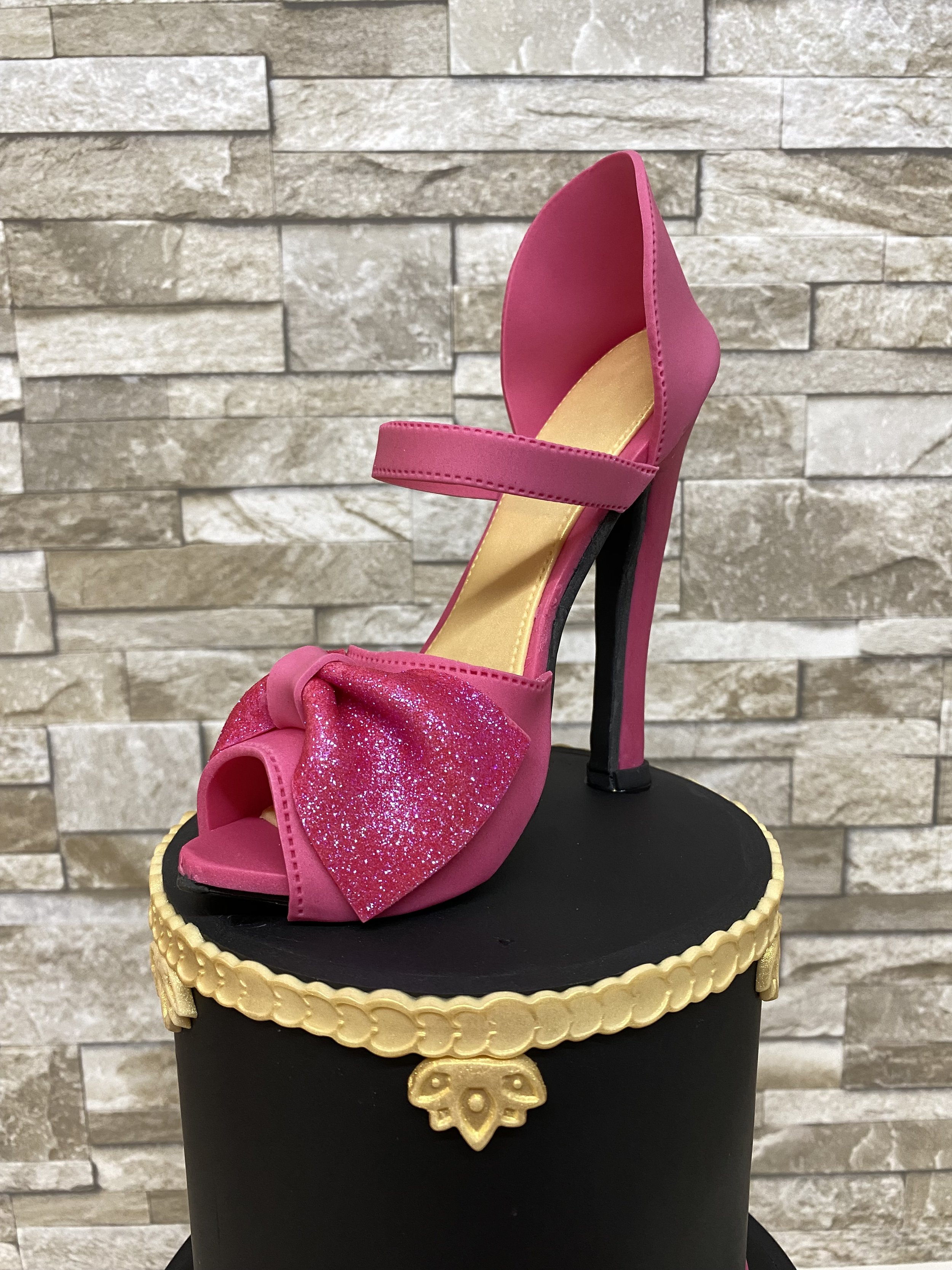 Fuchsia pink sugar stiletto with glitter bow on the toe seated on a black birthday cake with gold trim 