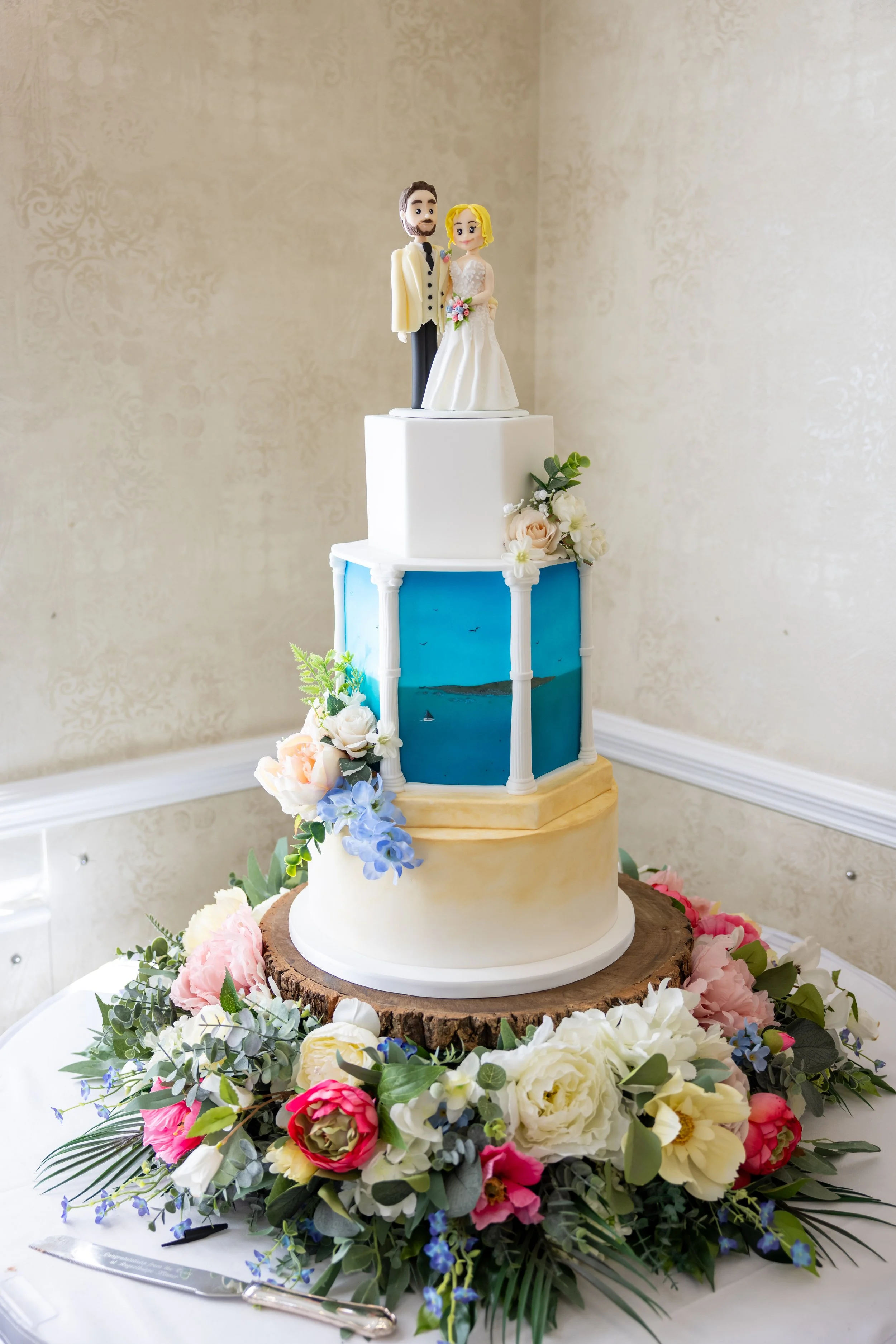 Three tiered wedding cake with a Mediterranean theme. Tall tier with blue sea view and greek style columns. Bunches of pastel flowers on ledges. Bride and groom cake topper on top. Seated on flower cake base