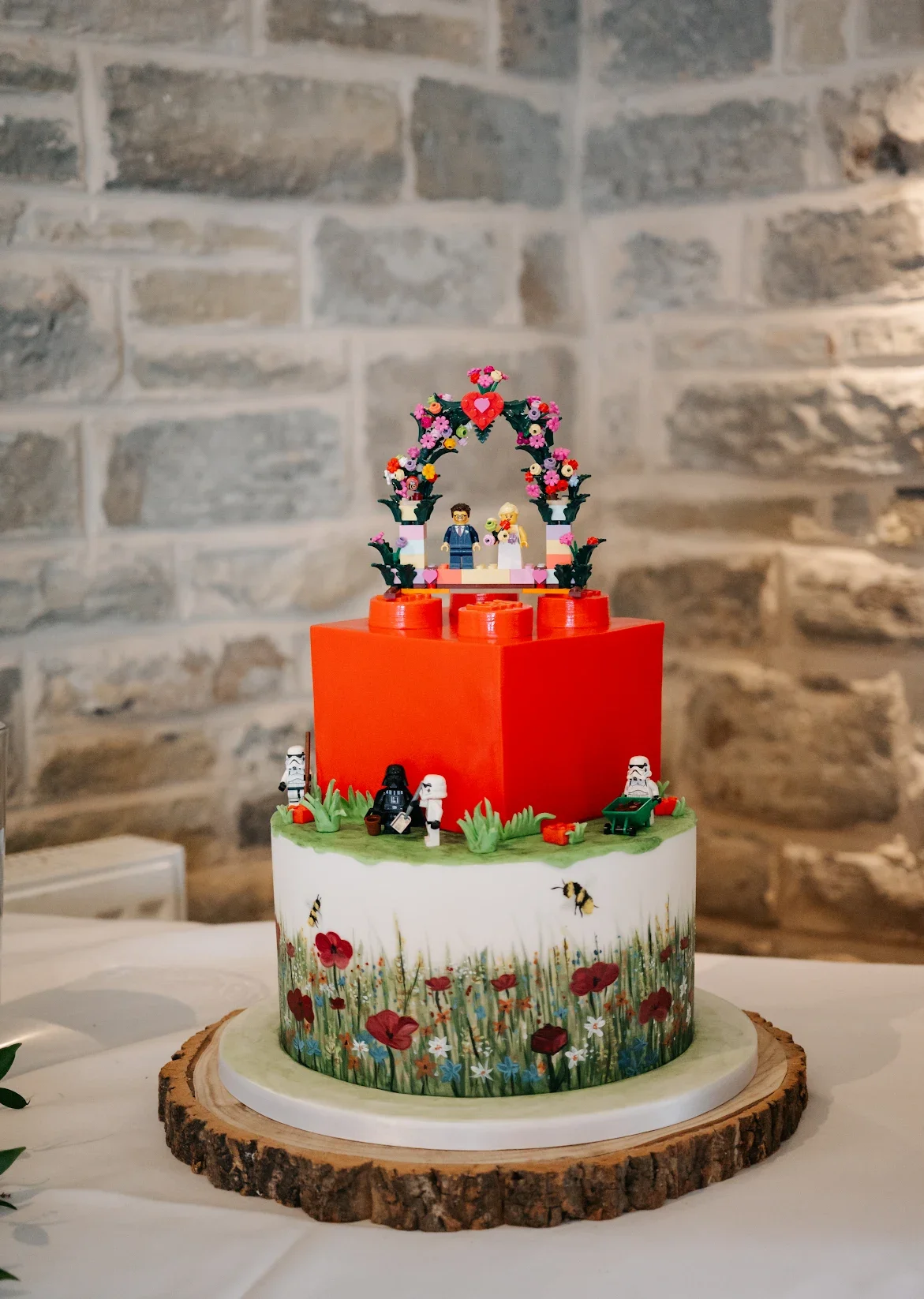 Two tier wedding cake featuring a hand painted wild meadow and red Lego brick, Lego figurines and Lego cake topper