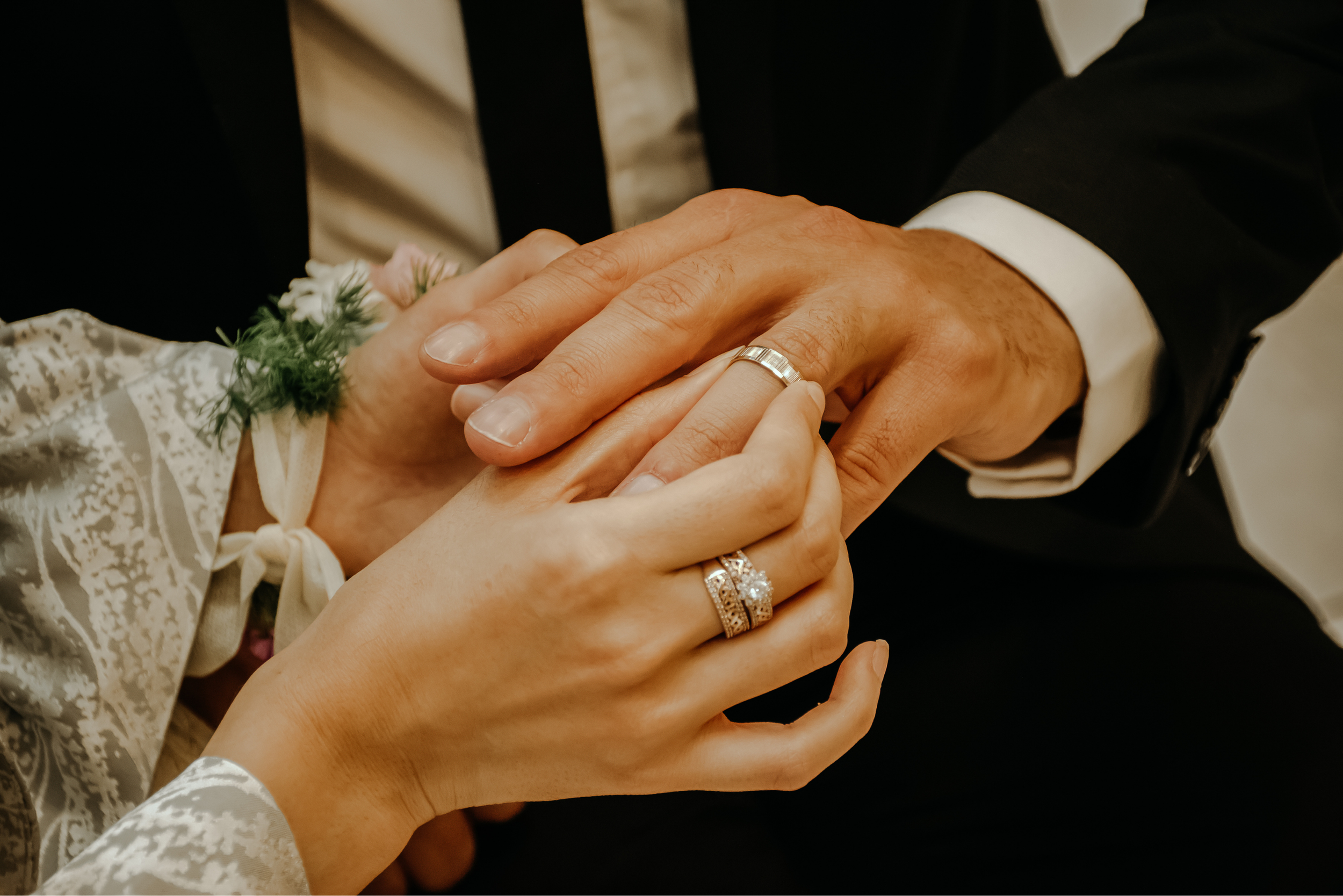 Close-up of a bride and groom holding hands, showing wedding rings; the bride's hand has a diamond engagement ring and a wedding band, and she wears a white lace dress with floral accents, while the groom wears a black suit with a white shirt and a striped tie.