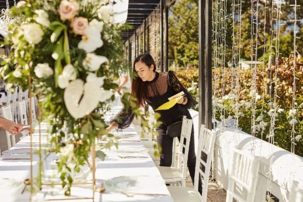 Woman setting up a decorated outdoor table for an event, with floral arrangements and string lights.