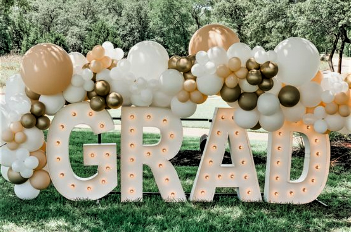 Decorative outdoor display with large illuminated letters spelling 'GRAZED' surrounded by balloons in white, gold, and beige colors, set on a grassy area with trees in the background.