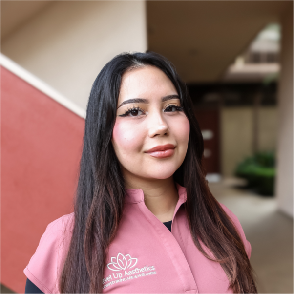 Young woman with long dark hair wearing pink uniform with 'Revive Up Aesthetics' logo, standing outdoors near a building.