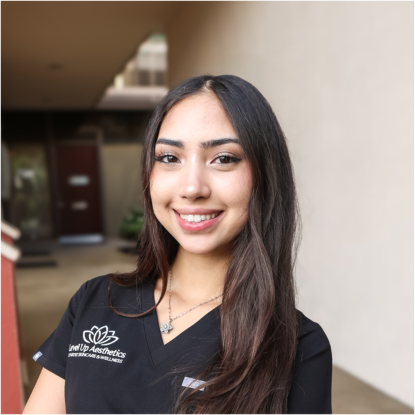 Young woman with long brown hair smiling outdoors, wearing black scrubs with a medical logo.