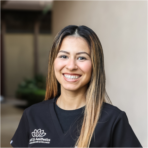 A smiling woman with long, light brown hair wearing a black uniform with the logo for Make Up Aesthetics on it, standing outdoors.