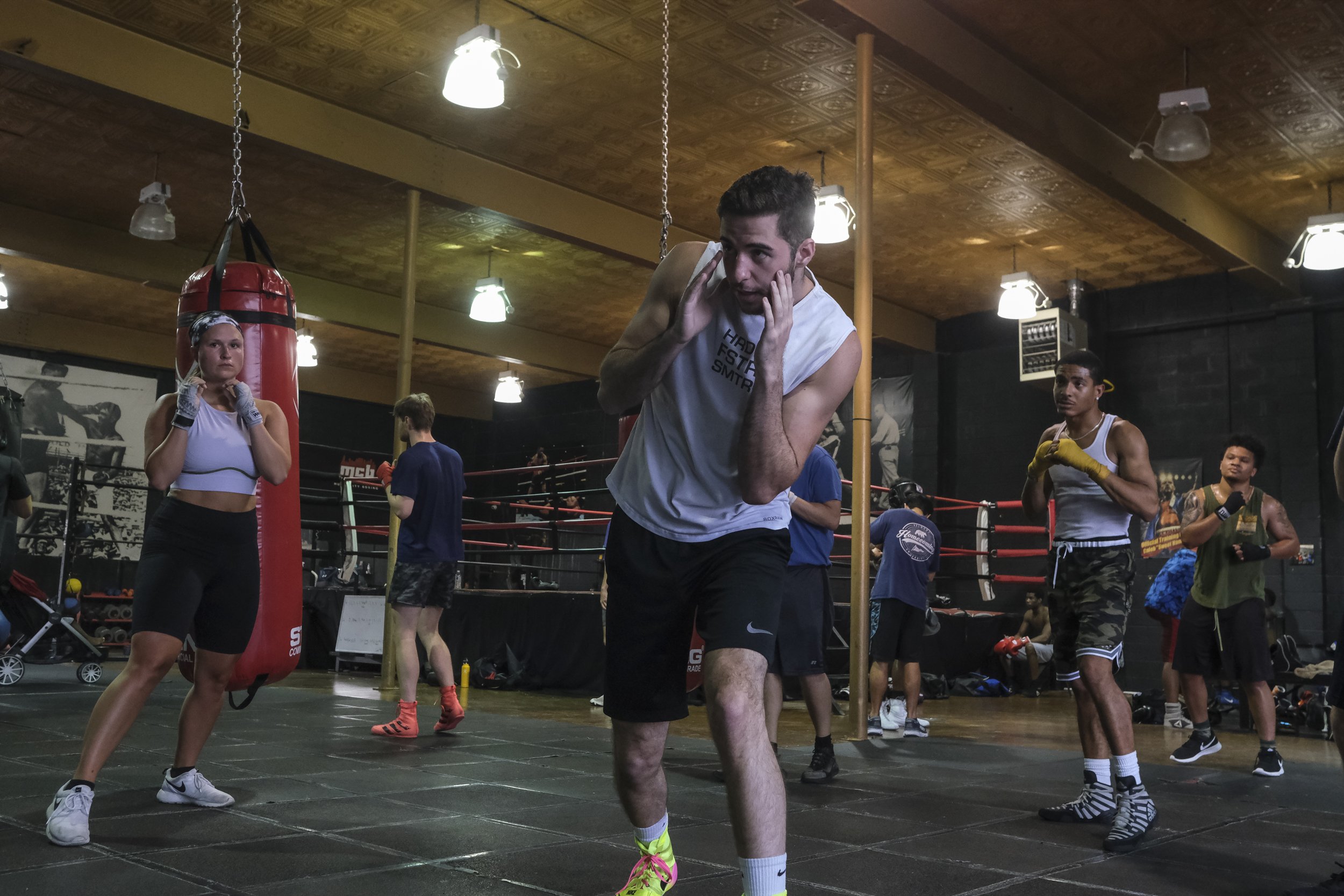 People in a boxing gym practicing and sparring with focus mitts and gloves, with a punching bag hanging from the ceiling.