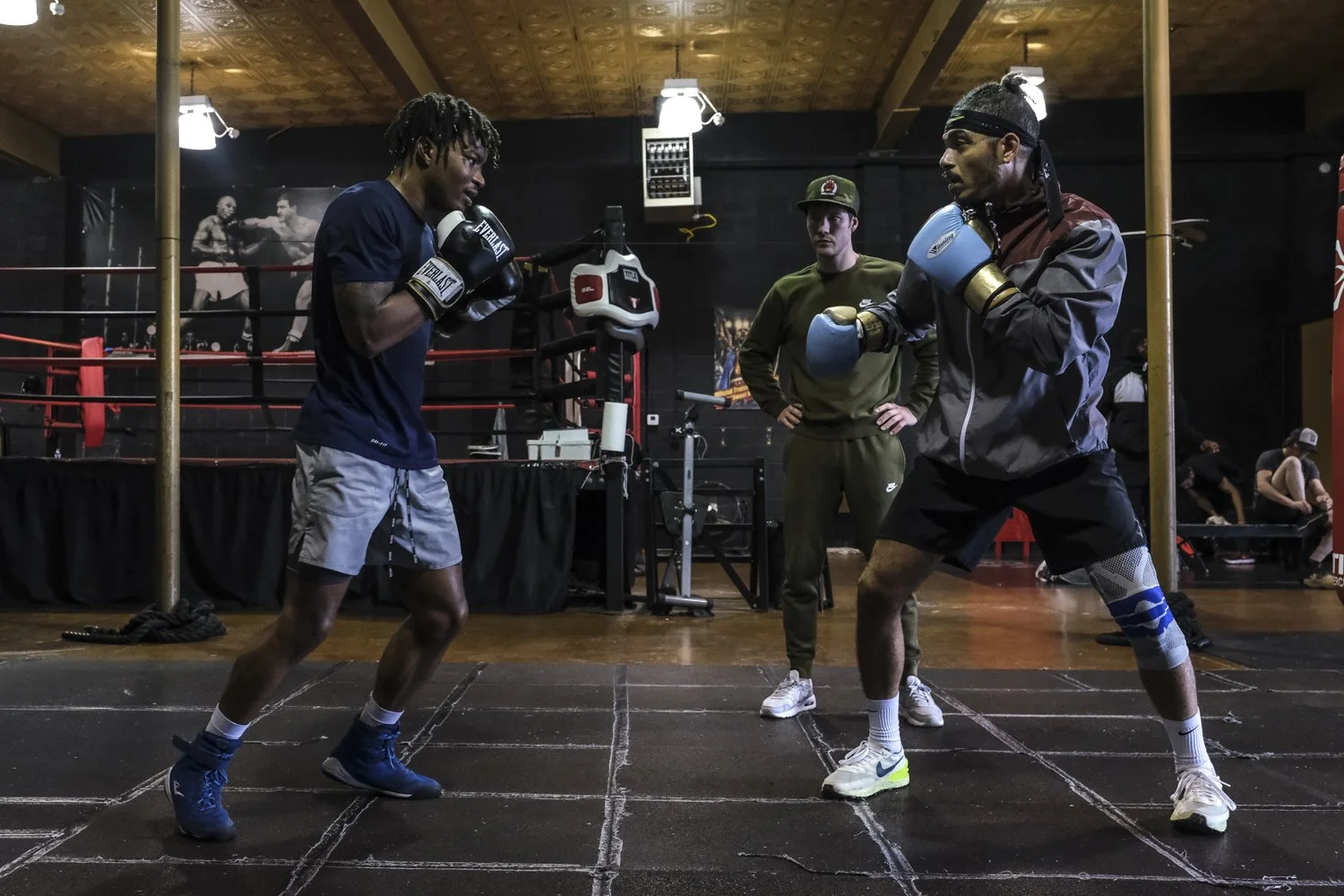 Two male boxers training in a boxing gym, one wearing blue gloves and the other with light blue gloves, with a coach standing behind them. The gym has a boxing ring in the background and a boxing poster on the wall.