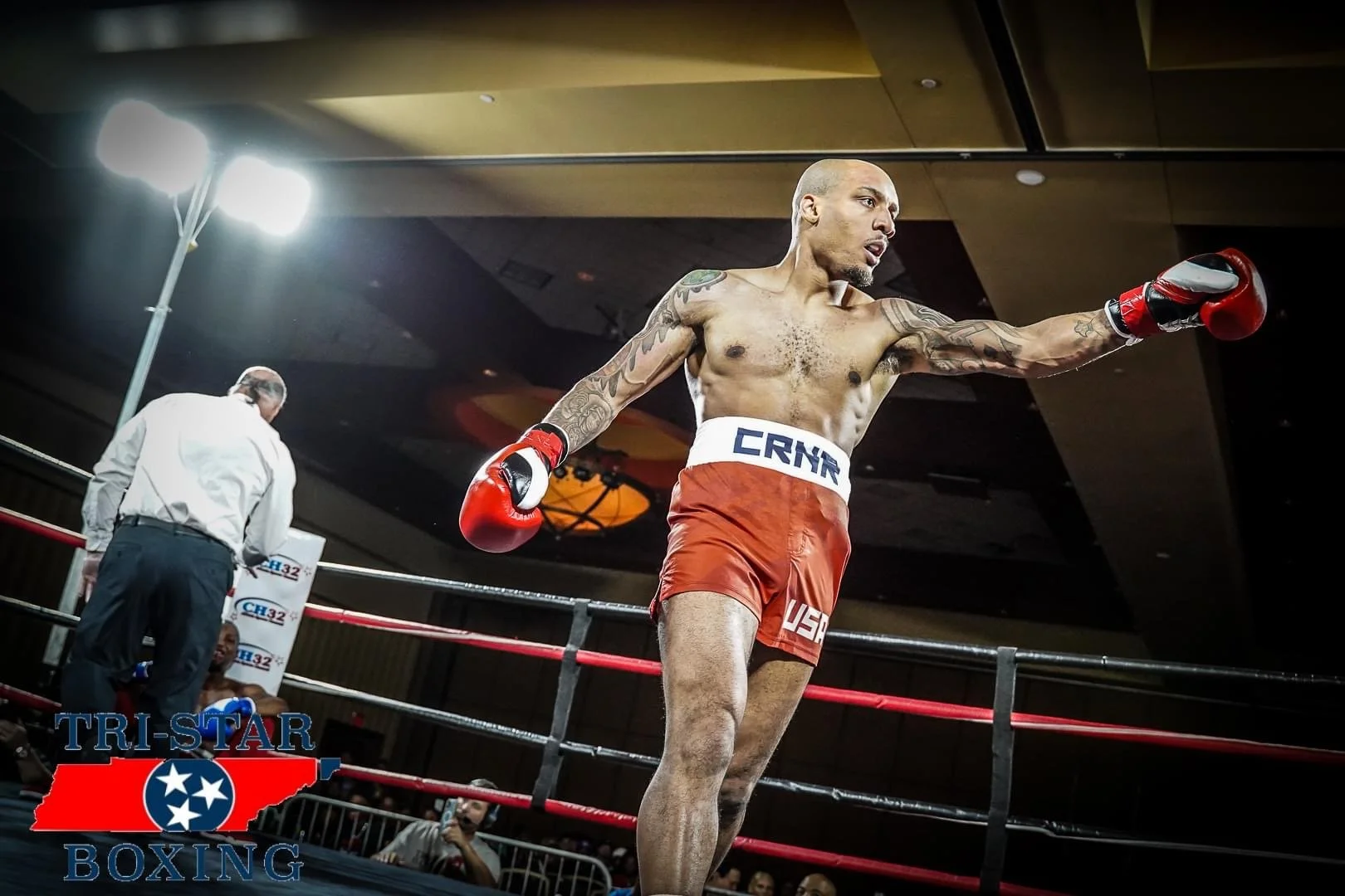 A boxer with tattoos wearing red boxing gloves and red shorts is practicing in a boxing ring. There is a referee in the background, and the TRISTAR Boxing logo is visible in the bottom left corner.