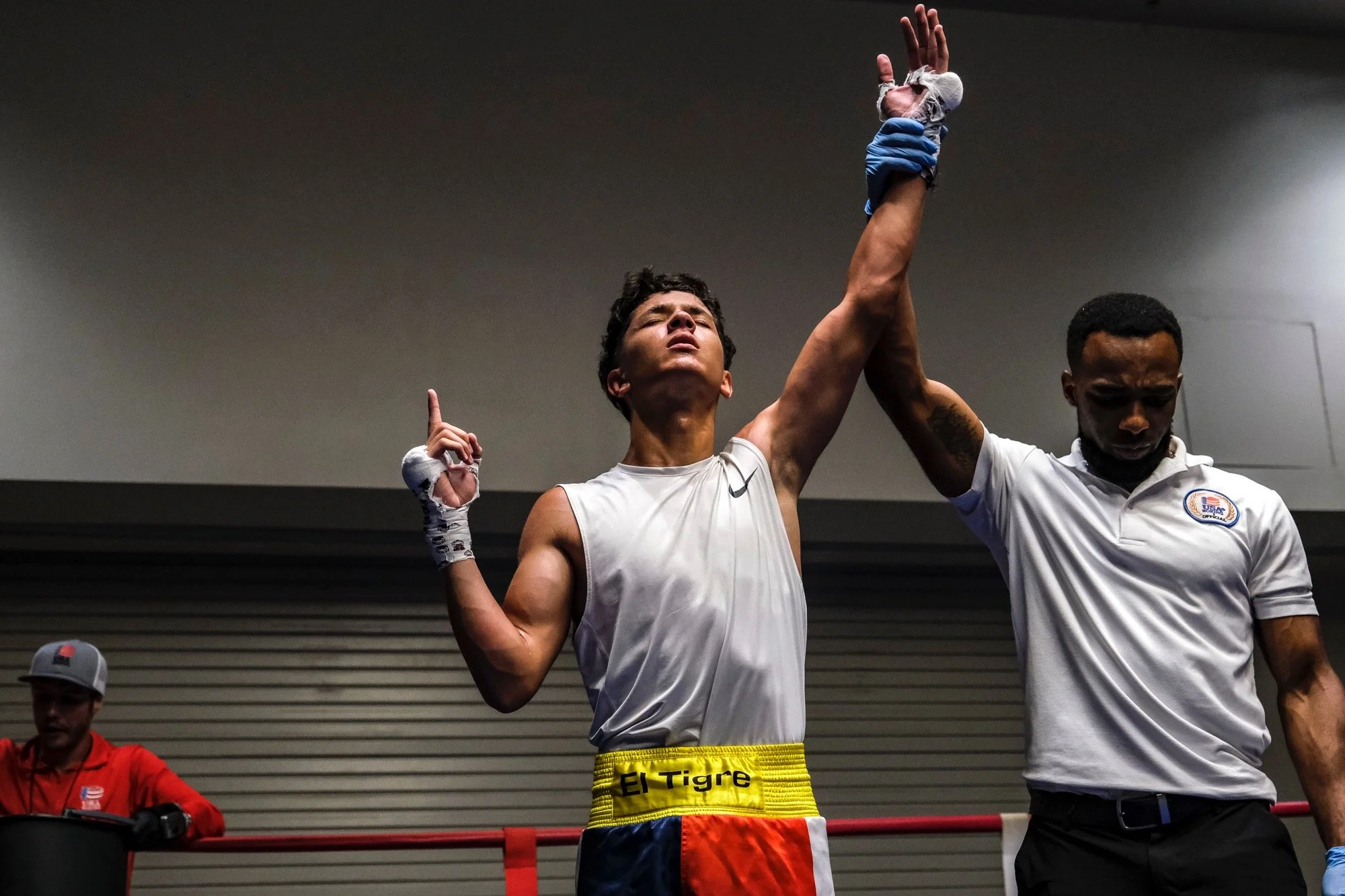 A young boxer with a muscular build and curly hair is in a boxing ring, eyes closed, head tilted back, raising his left hand, which is held by a referee. The boxer is wearing a gray sleeveless shirt, multicolored boxing shorts with an 'El Tigre' wais