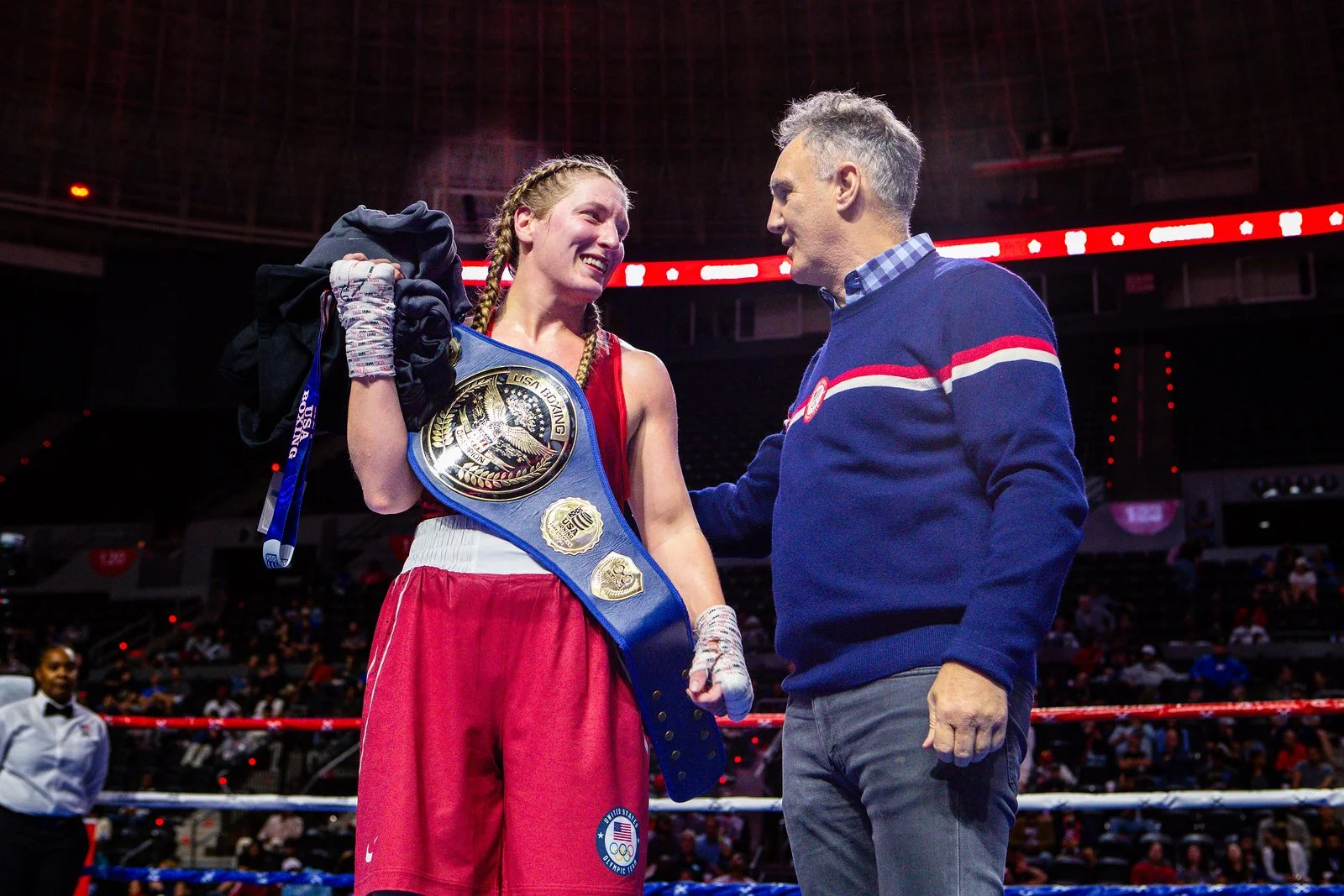 A female boxer with braided hair and a red and white uniform, holding a championship belt, smiling at a man in a blue sweater, inside a boxing ring with a crowd in the background.