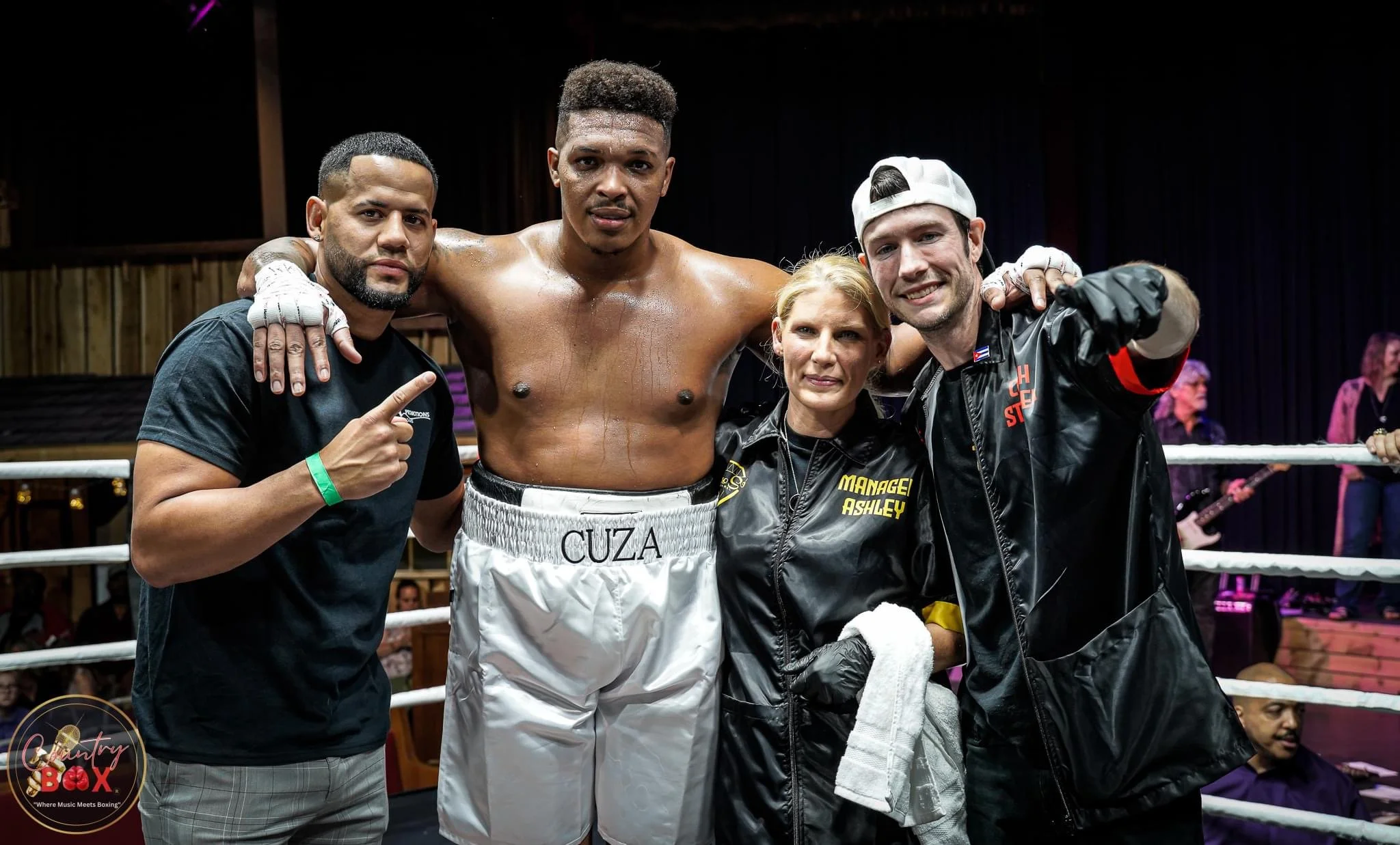 Boxer and team celebrating in the boxing ring after a match, with a woman and two men, all smiling, with the boxer shirtless and wearing gloves.