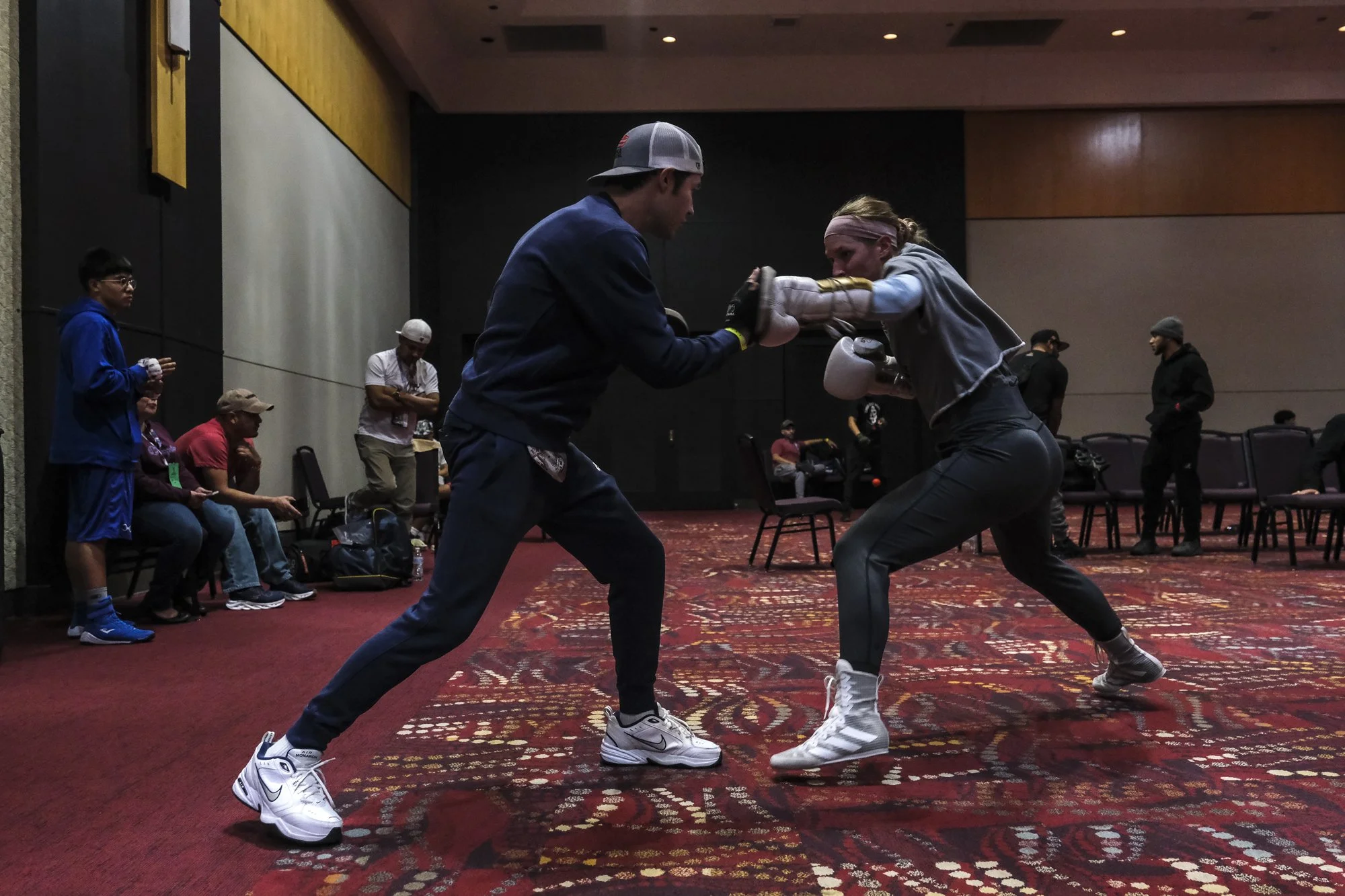 Two people practicing boxing in an indoor training area, with onlookers seated and standing in the background.